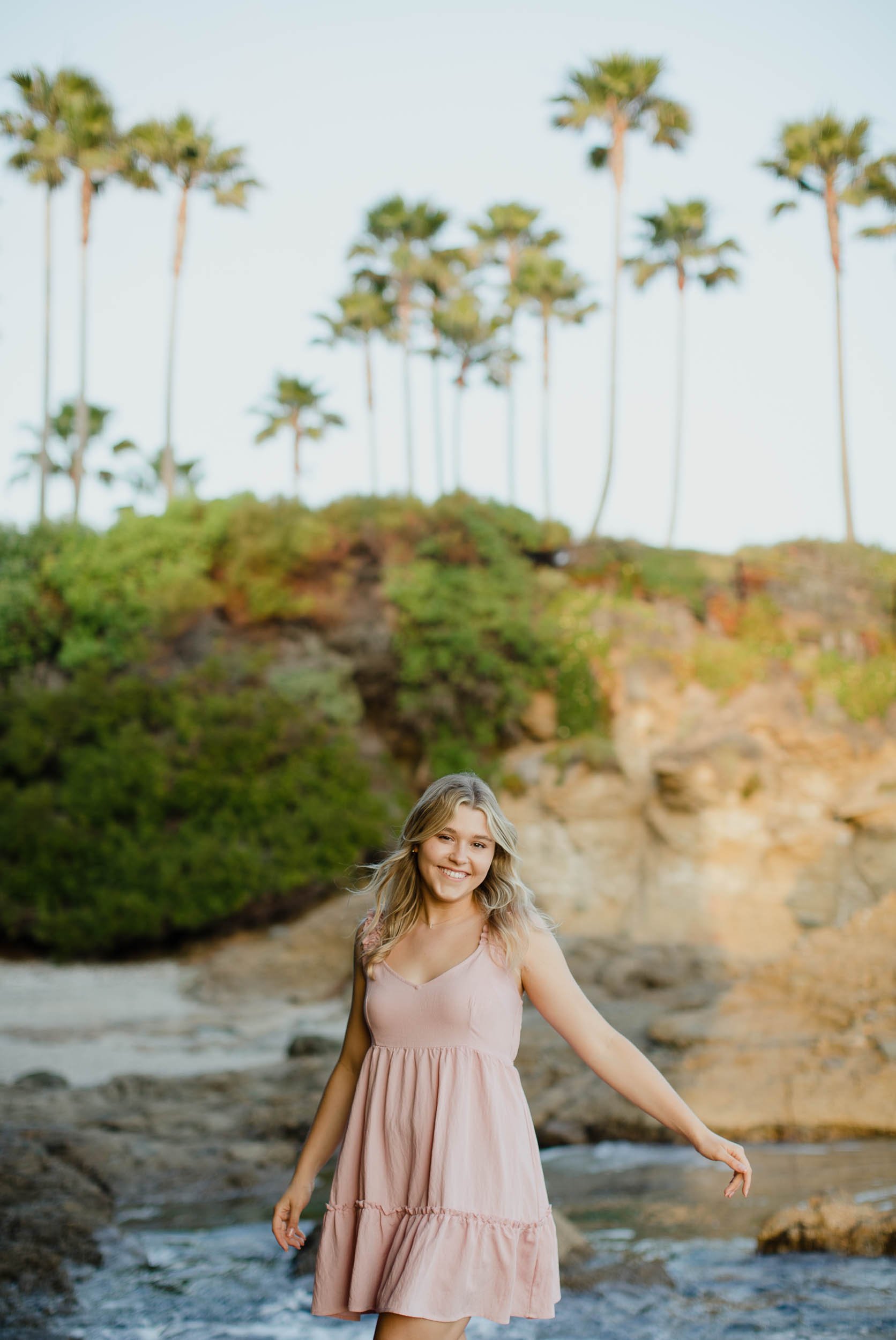 High school senior portrait girl in a pink dress standing near rocky shoreline with palm trees in the background.