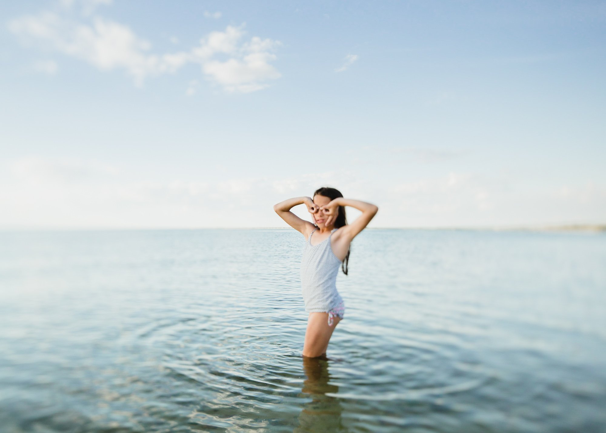 Orange County Child and Family Photography. Girl in swimsuit standing in shallow ocean water, posing playfully with hands on her face.