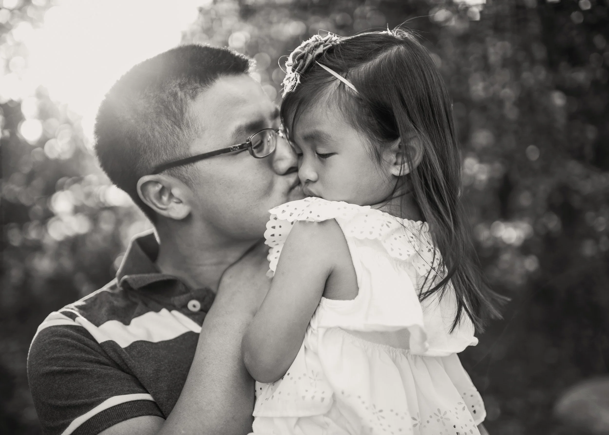 A black and white photo of a father wearing glasses kissing a young girl on the forehead. The girl is wearing a white dress and has a flower headband. They are surrounded by a blurred outdoor background.