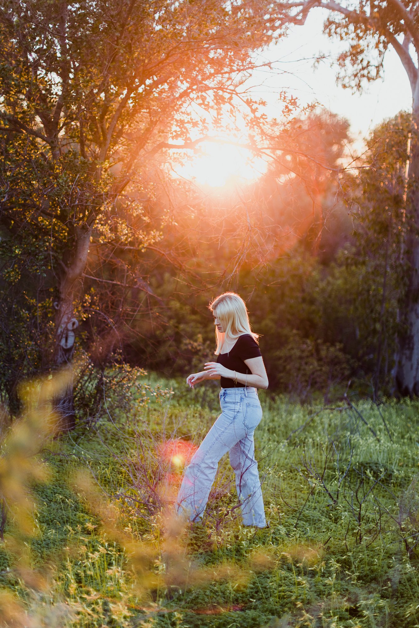 A person with blonde hair walking through a sunlit forest clearing, wearing a black top and light jeans. Sunlight filters through the trees, creating a warm glow.