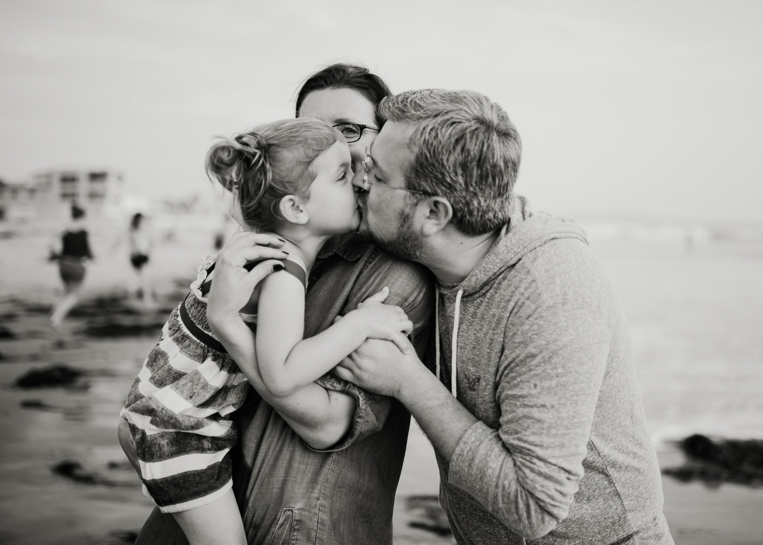 Orange County Child and Family Photography. A black and white photo of a family at the beach. A child is being embraced and kissed by two adults. The background shows a sandy beach with blurry figures and buildings.