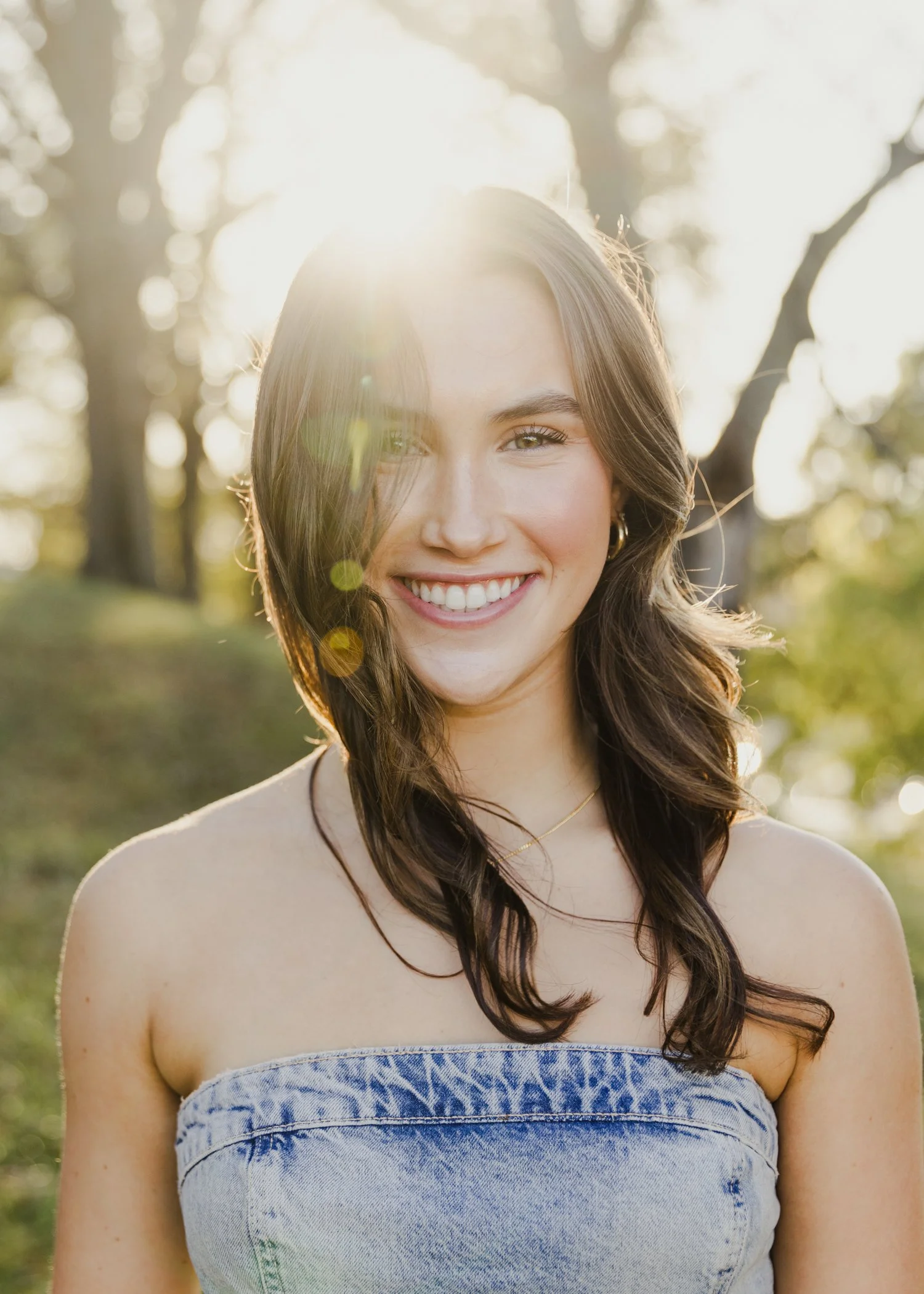 High school senior portrait girl smiling outdoors with sunlight filtering through trees, wearing a denim tube top.