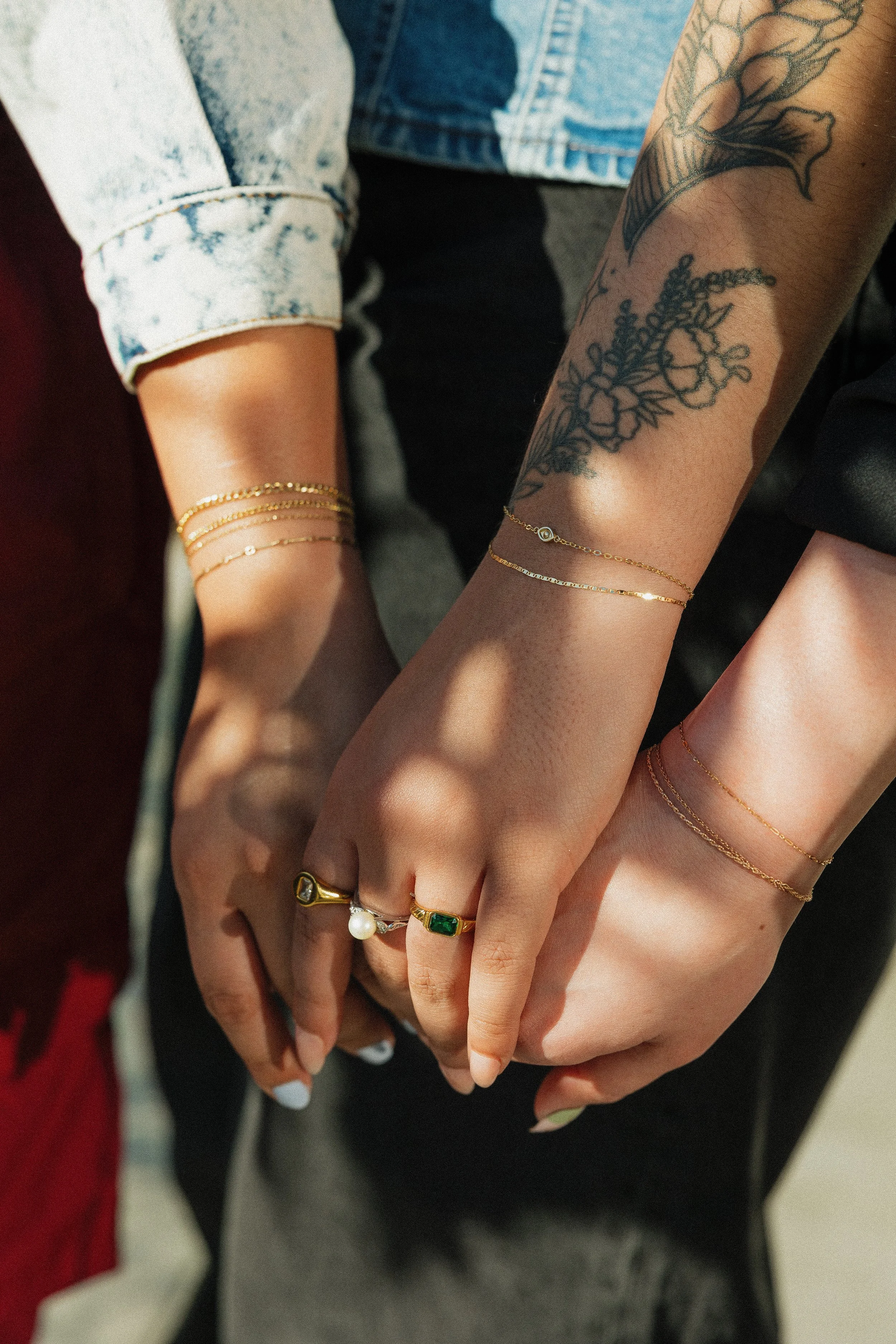 Close-up of two hands gently clasped, showing various gold and gemstone rings, and delicate gold bracelets. One hand features a black floral tattoo, and the other has a tattoo of flowers and leaves. The background is blurred, with part of denim cloth