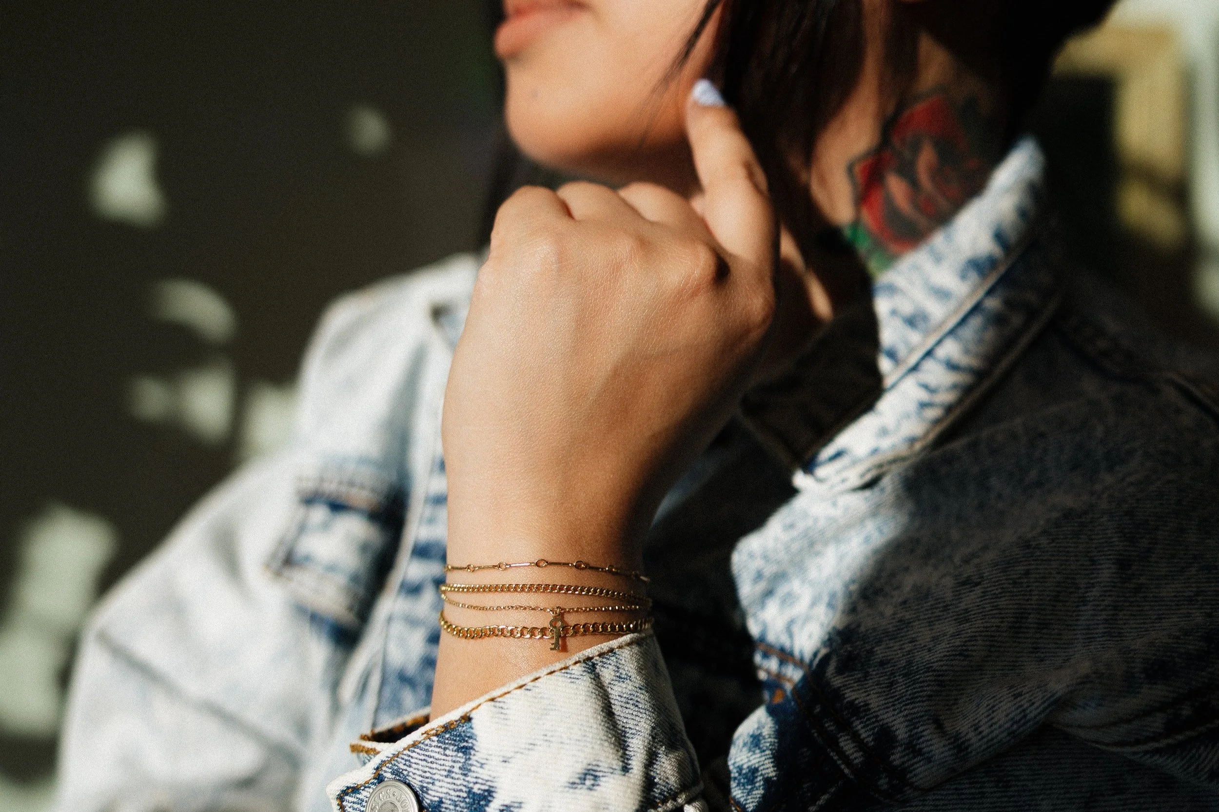 Close-up of a woman resting her chin on her hand, wearing multiple gold bracelets, denim jacket, and showing colorful tattoos on her neck and face.