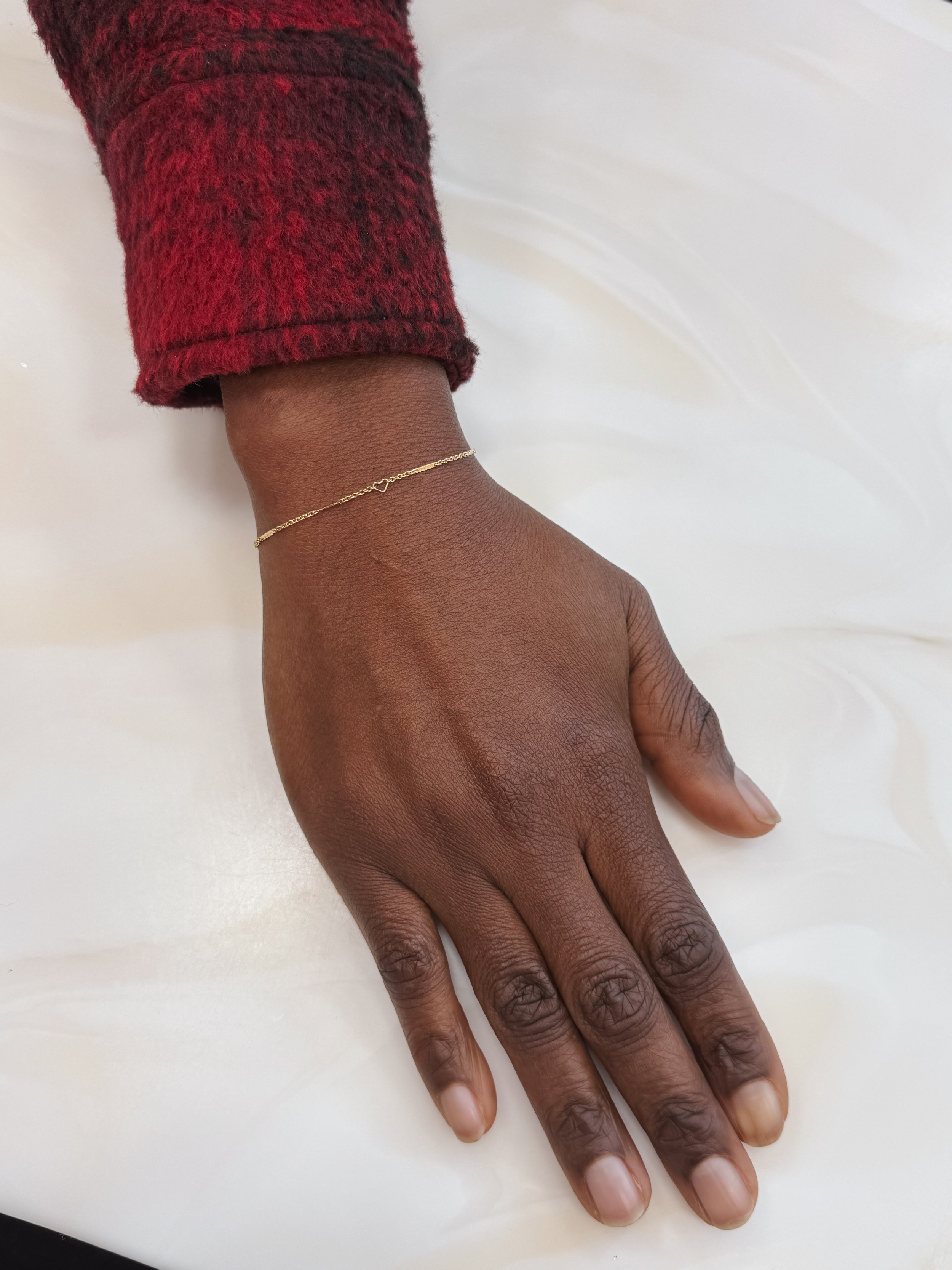 Close-up of a person's hand resting on a white surface, wearing a delicate solid 14k gold permanent bracelet and a red and black sweater sleeve.