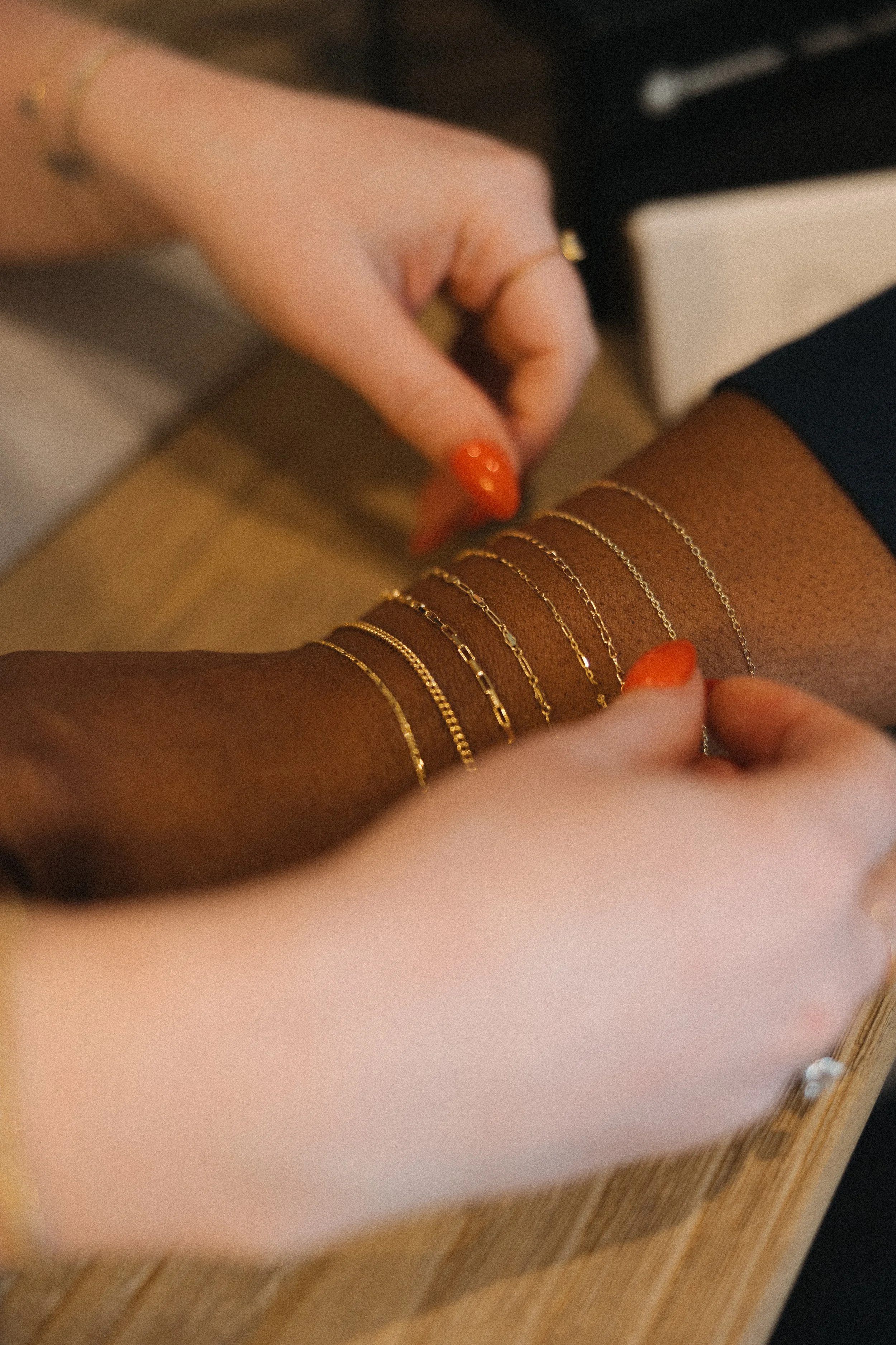 Close-up of a person's arm with multiple gold bracelets, with a hand adjusting them, and another hand with orange nail polish holding the arm.