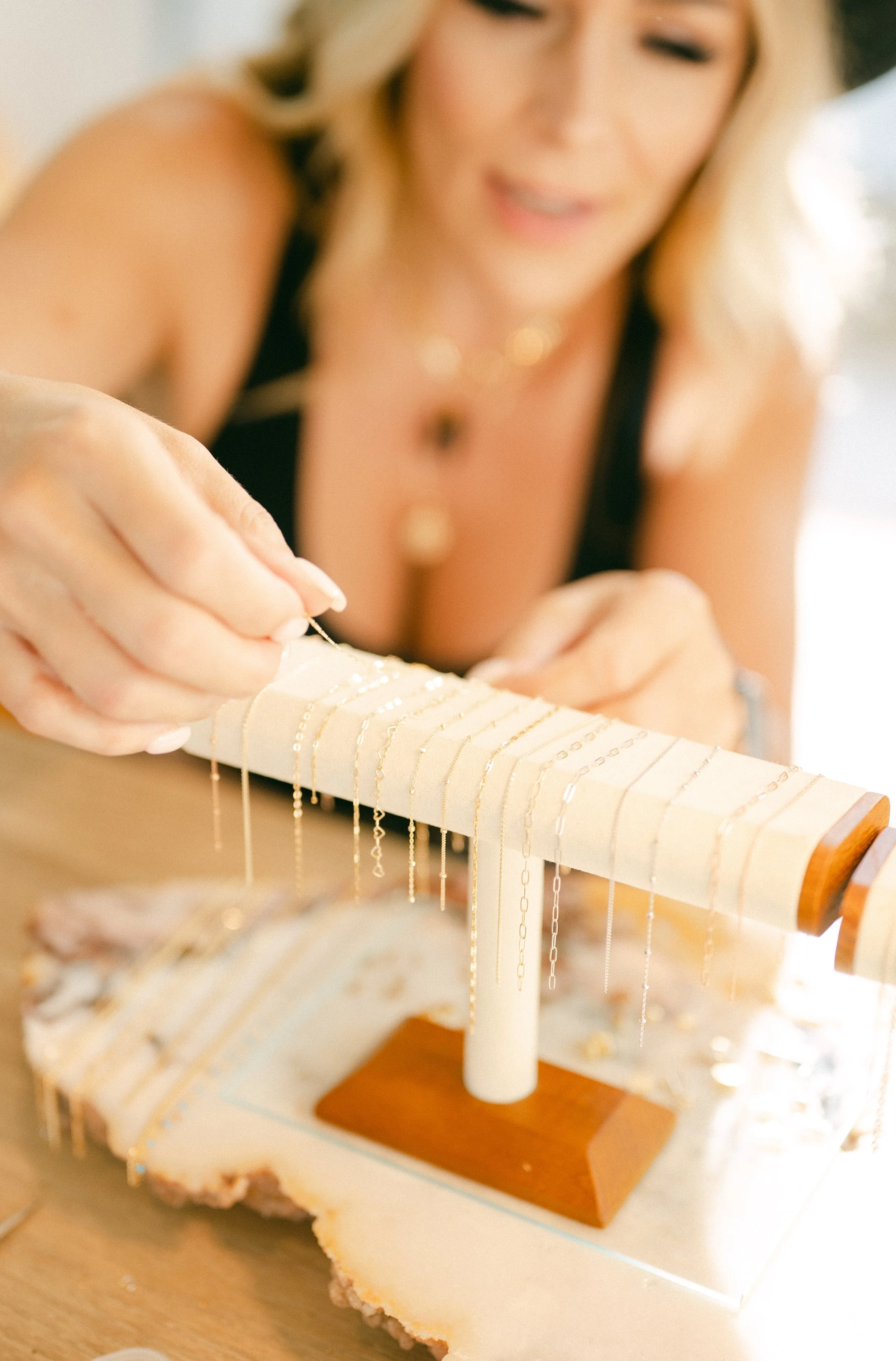 A woman with blonde hair, wearing a black top and jewelry, is selecting a delicate gold necklace from a jewelry stand.