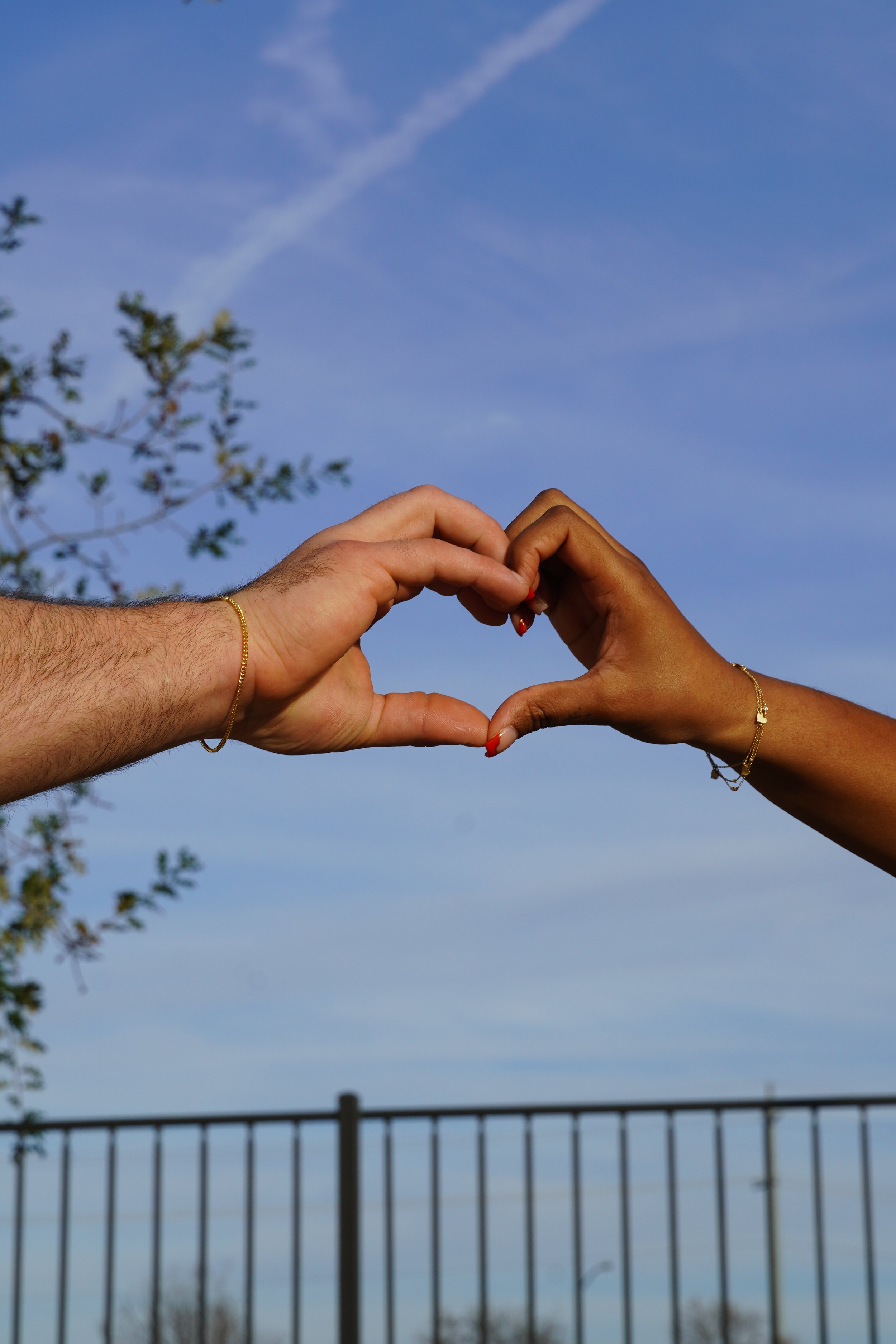 Two people form a heart shape with their hands with permanent bracelets against a blue sky with clouds, trees, and a fence in the background.