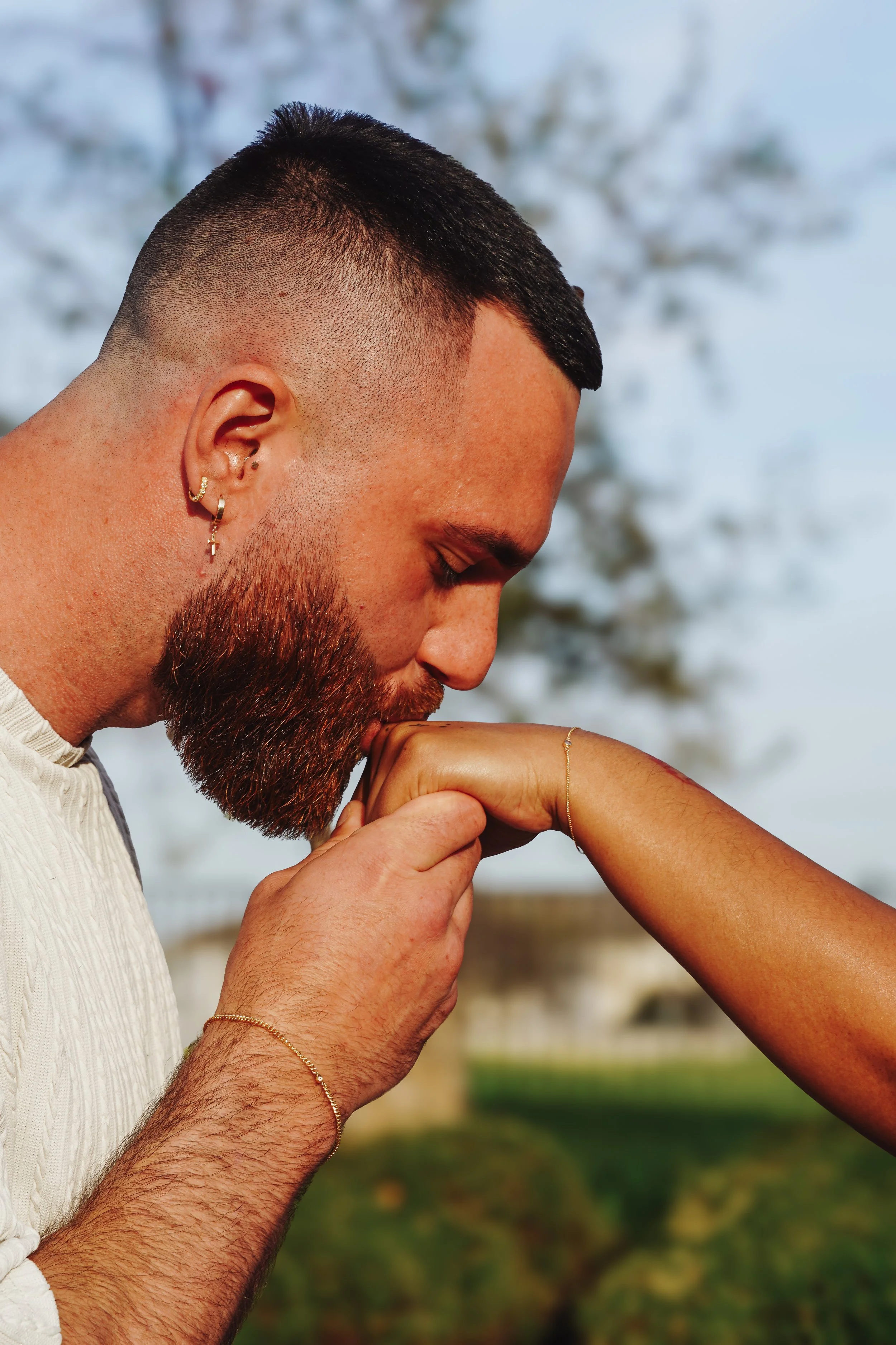 A man with a short beard and earrings kisses a woman's hand wearing permanent bracelets.