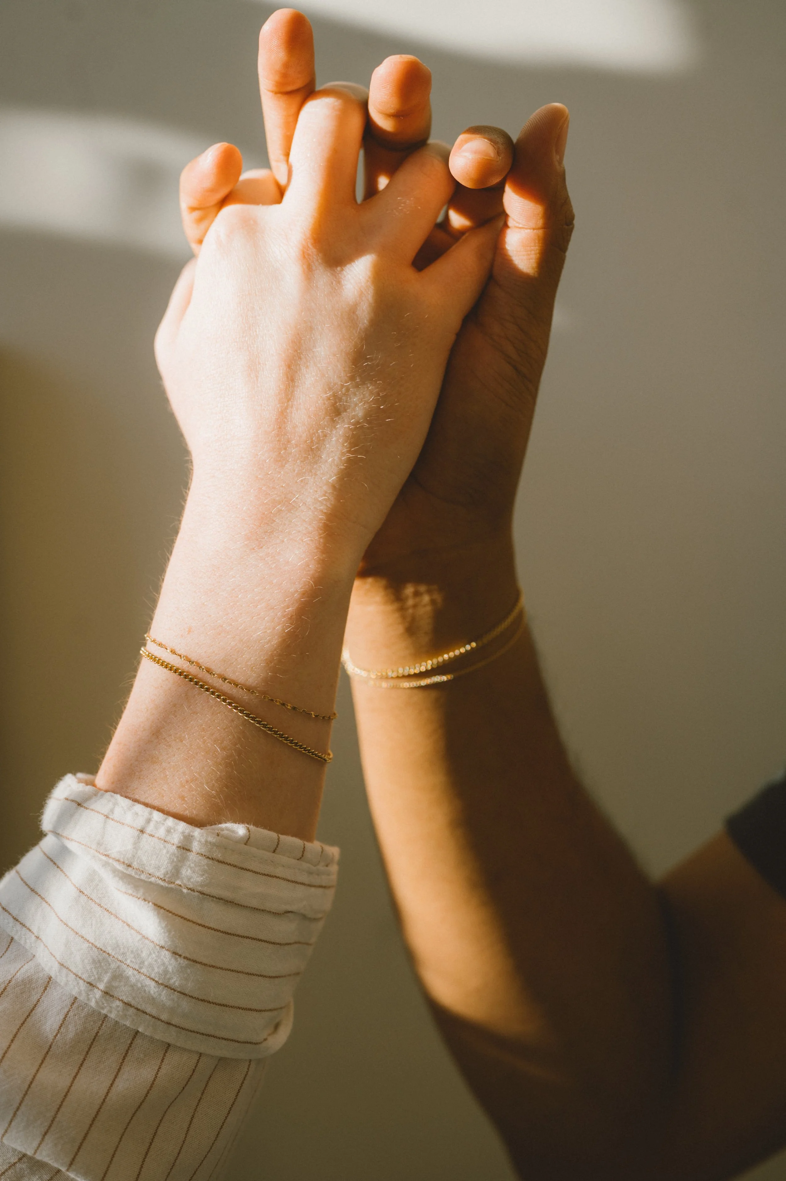 Two hands gently clasped, with sunlight casting shadows, wearing delicate gold bracelets and one person wearing a striped shirt sleeve.