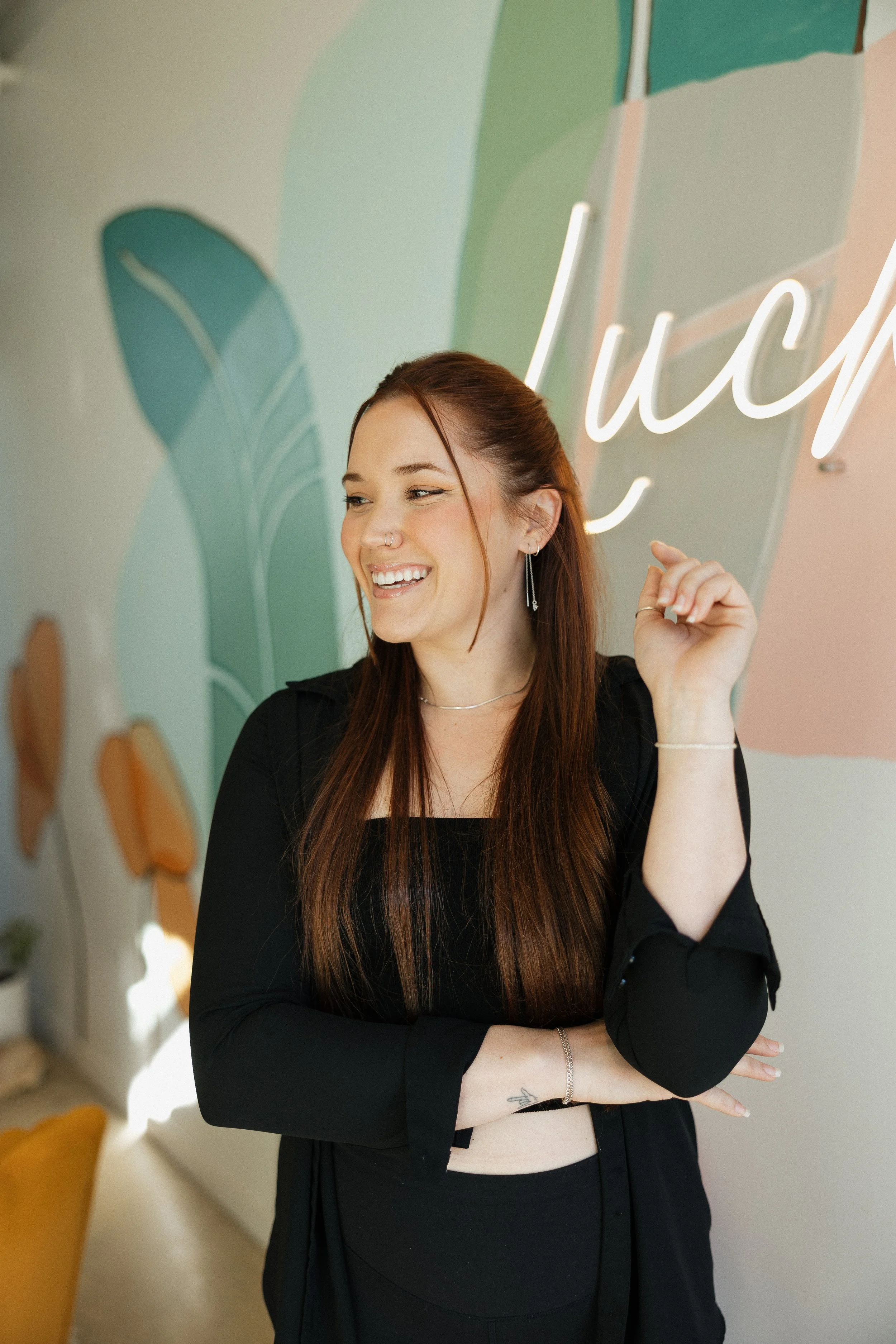 A young woman with long red hair, wearing black clothing and jewelry, smiles and crosses her arms while standing in front of a colorful wall with the word 'lucky' written on it.