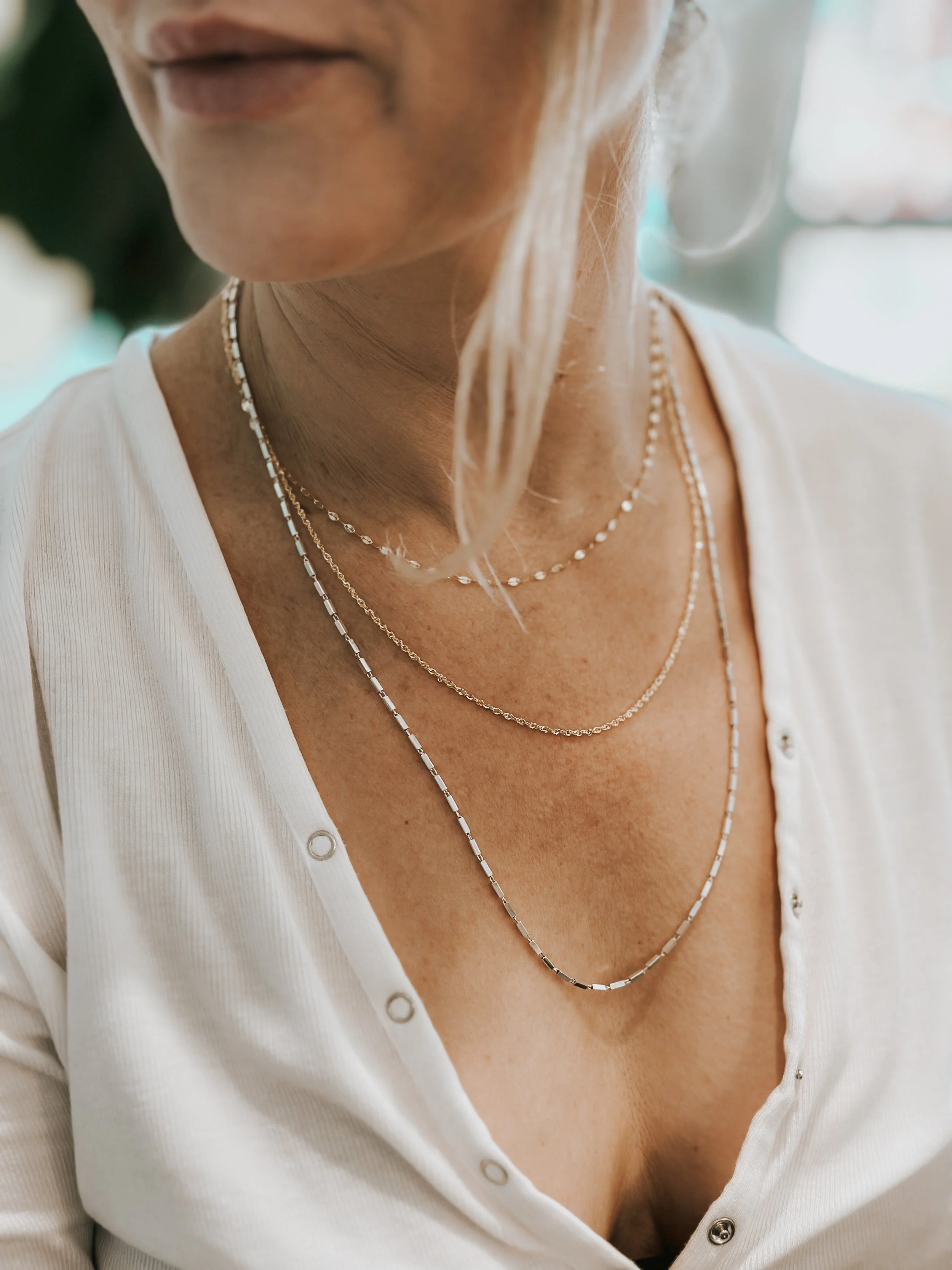 Close-up of a woman wearing layered delicate necklaces with a white shirt that has a few buttons open at the top.