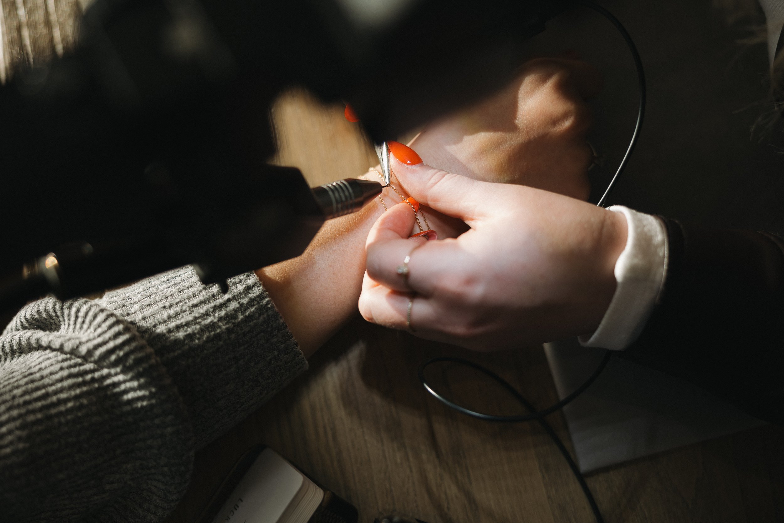 A jewelry artist engraves a necklace on a workbench, wearing a gray sweater and using a dental handpiece.