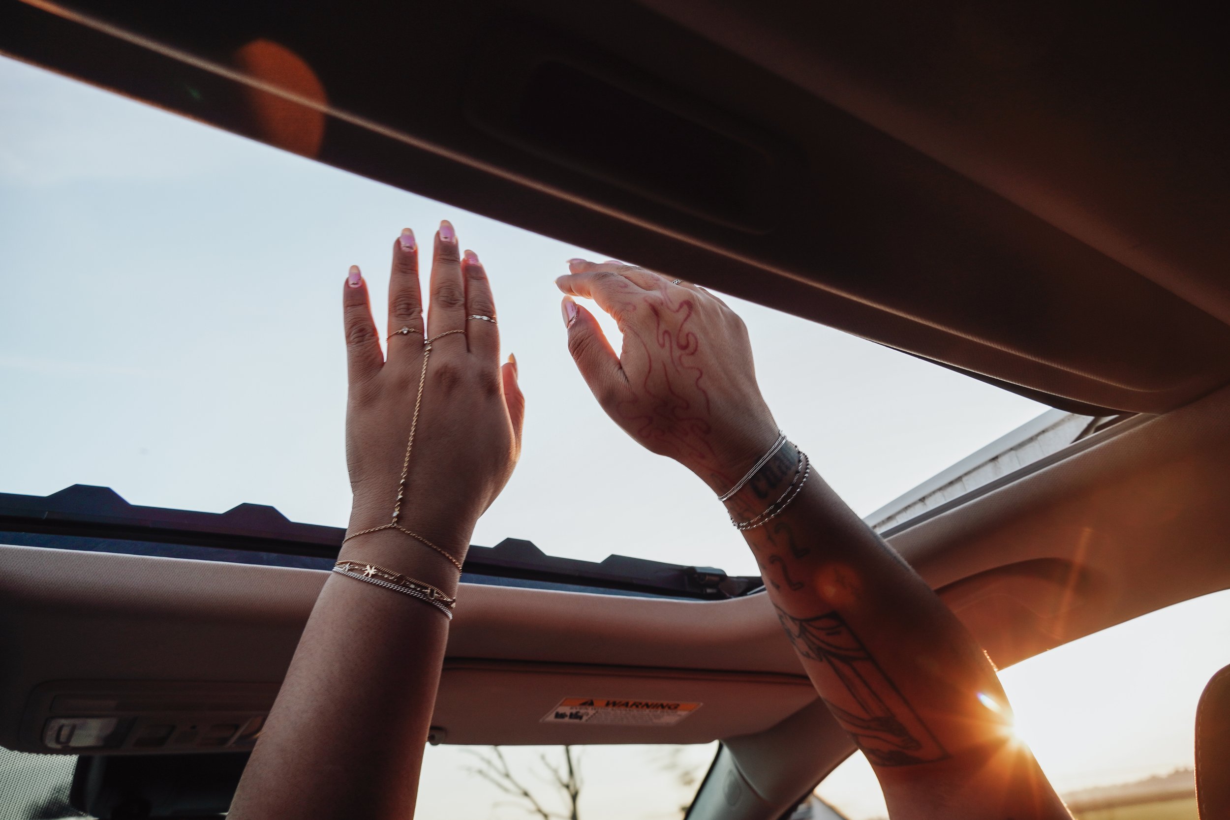 Two hands, one with permanent jewelry and tattoos, reaching up through the open sunroof of a car toward the sky during sunset.