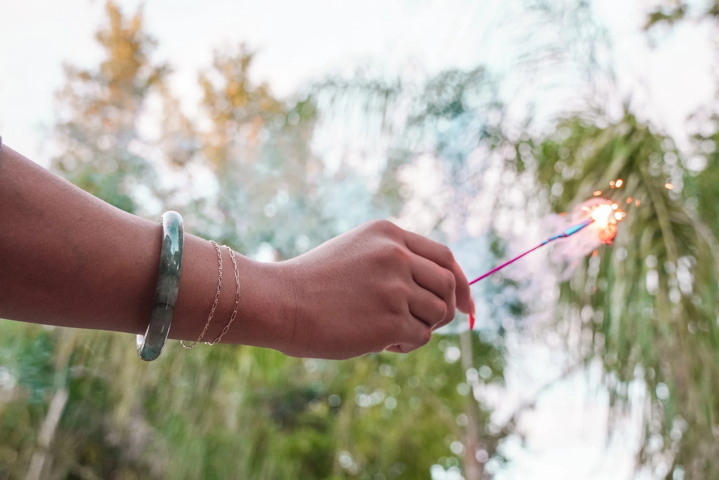 A person wearing amazing permanent jewelry from Lucky Gold holding a lit sparkler outdoors during daytime, with blurred trees in the background.