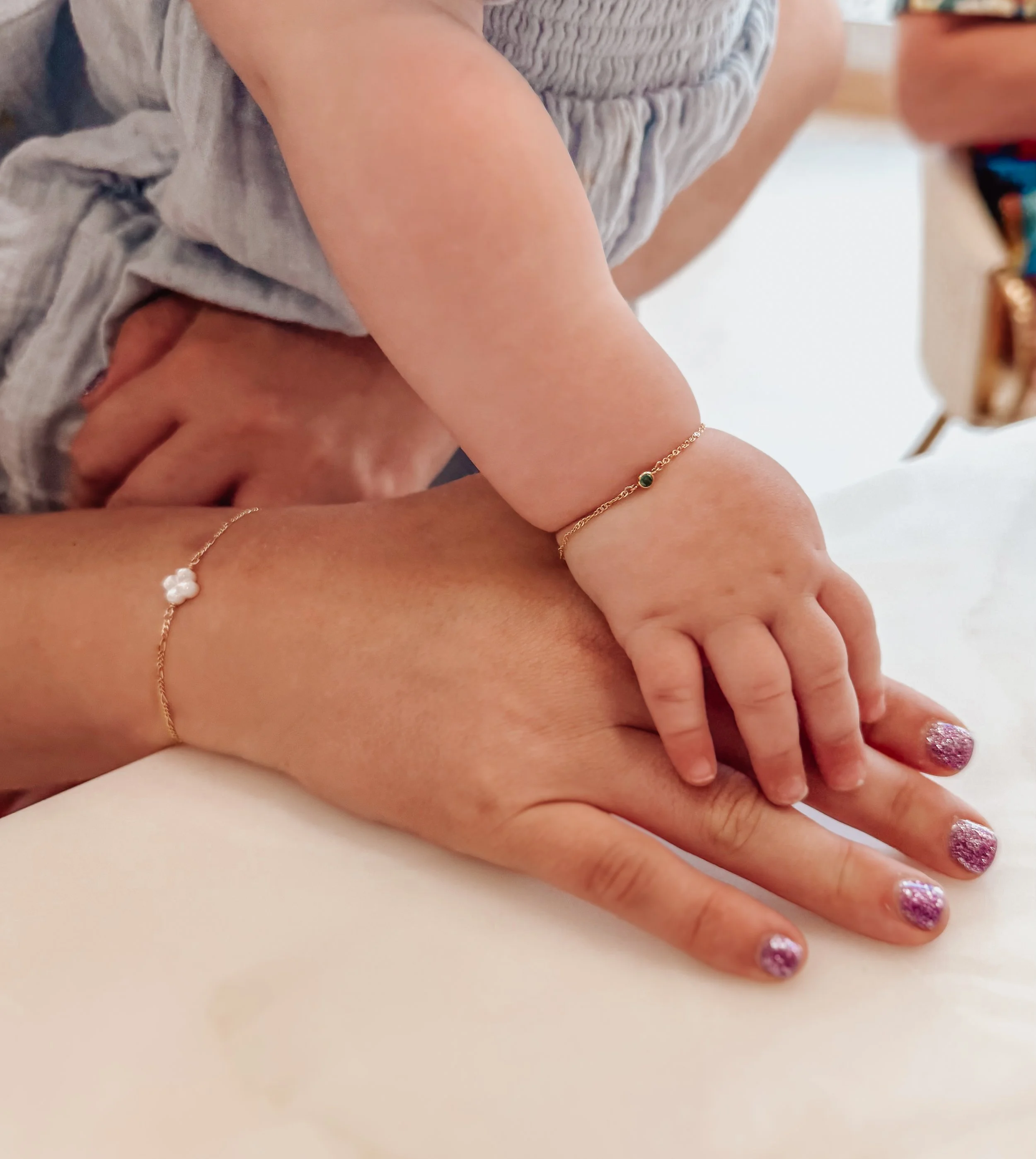 Close-up of a baby's hand gently resting on an adult's hand, both wearing delicate 14k permanent bracelets.