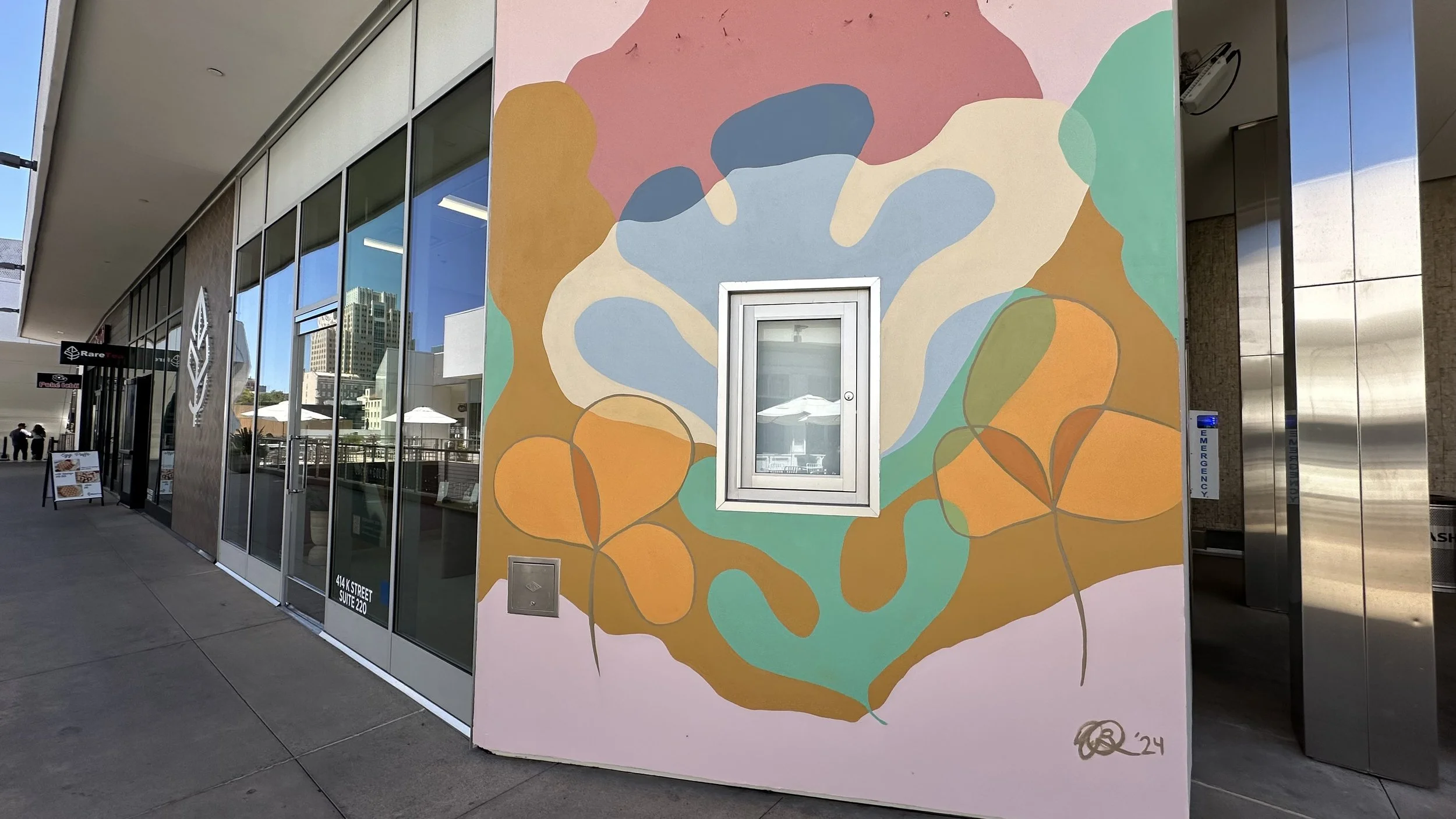 A colorful abstract mural painted on a building exterior, featuring organic shapes in pink, blue, orange, green, and beige with a small window and a metallic mail slot.