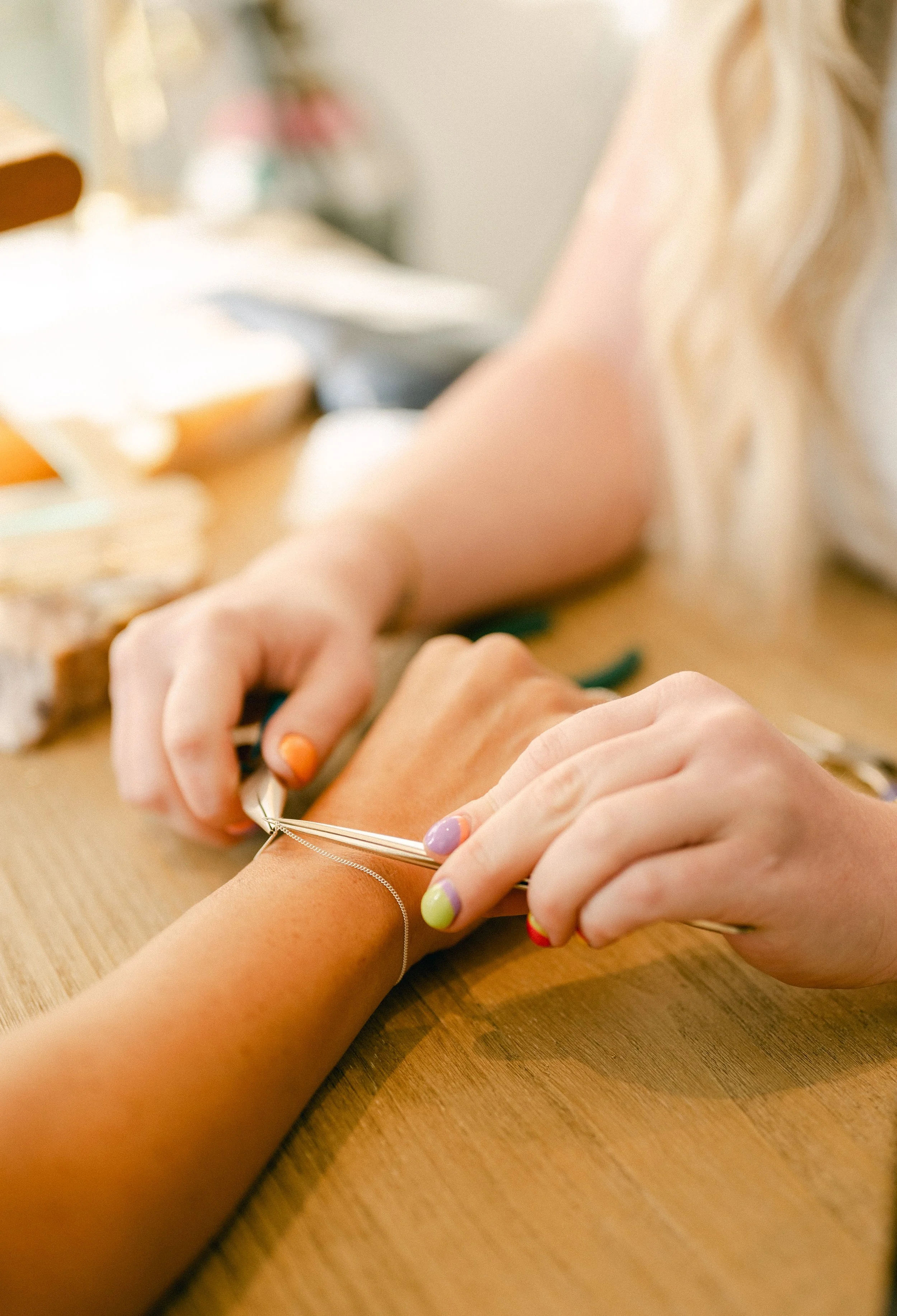 A person wearing colorful painted nails is putting a silver chain on another person's wrist, which already has a delicate jewelry chain.