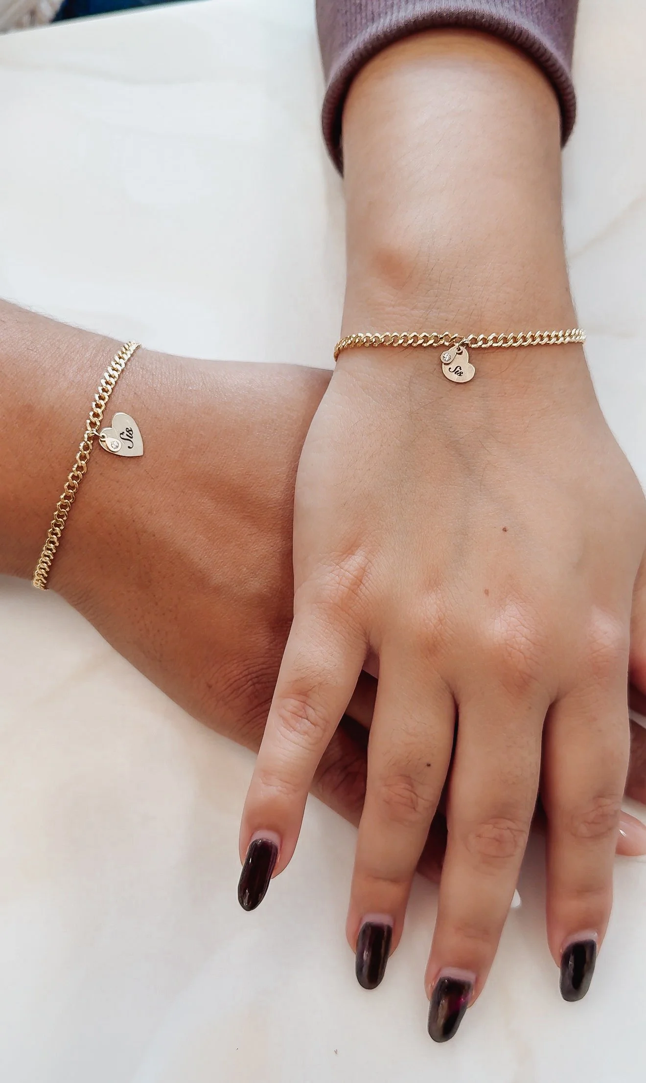 Close-up of two hands with matching gold heart-shaped permanent bracelets engraved with the word 'Sue' and decorated with a small gemstone.