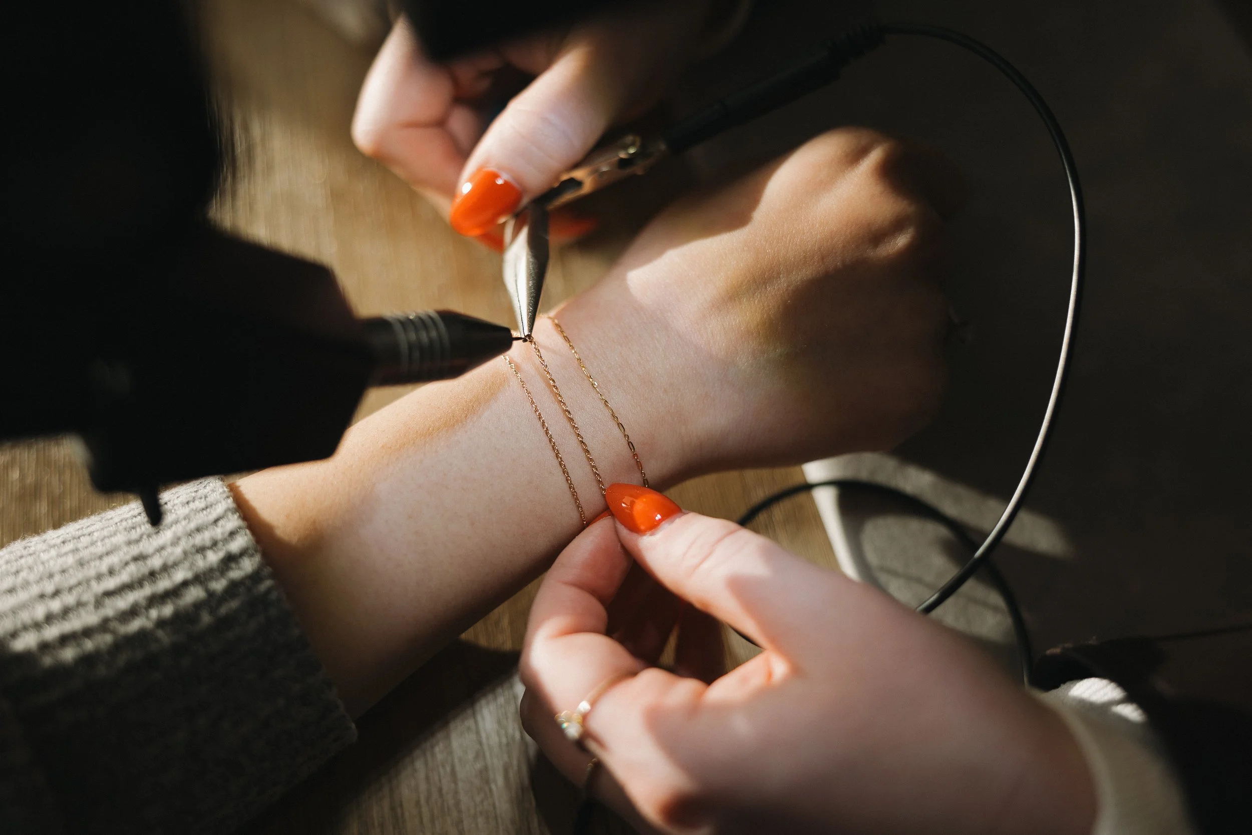 Person is creating or repairing a delicate gold necklace with a soldering device, with tools and jewelry on a wooden surface.