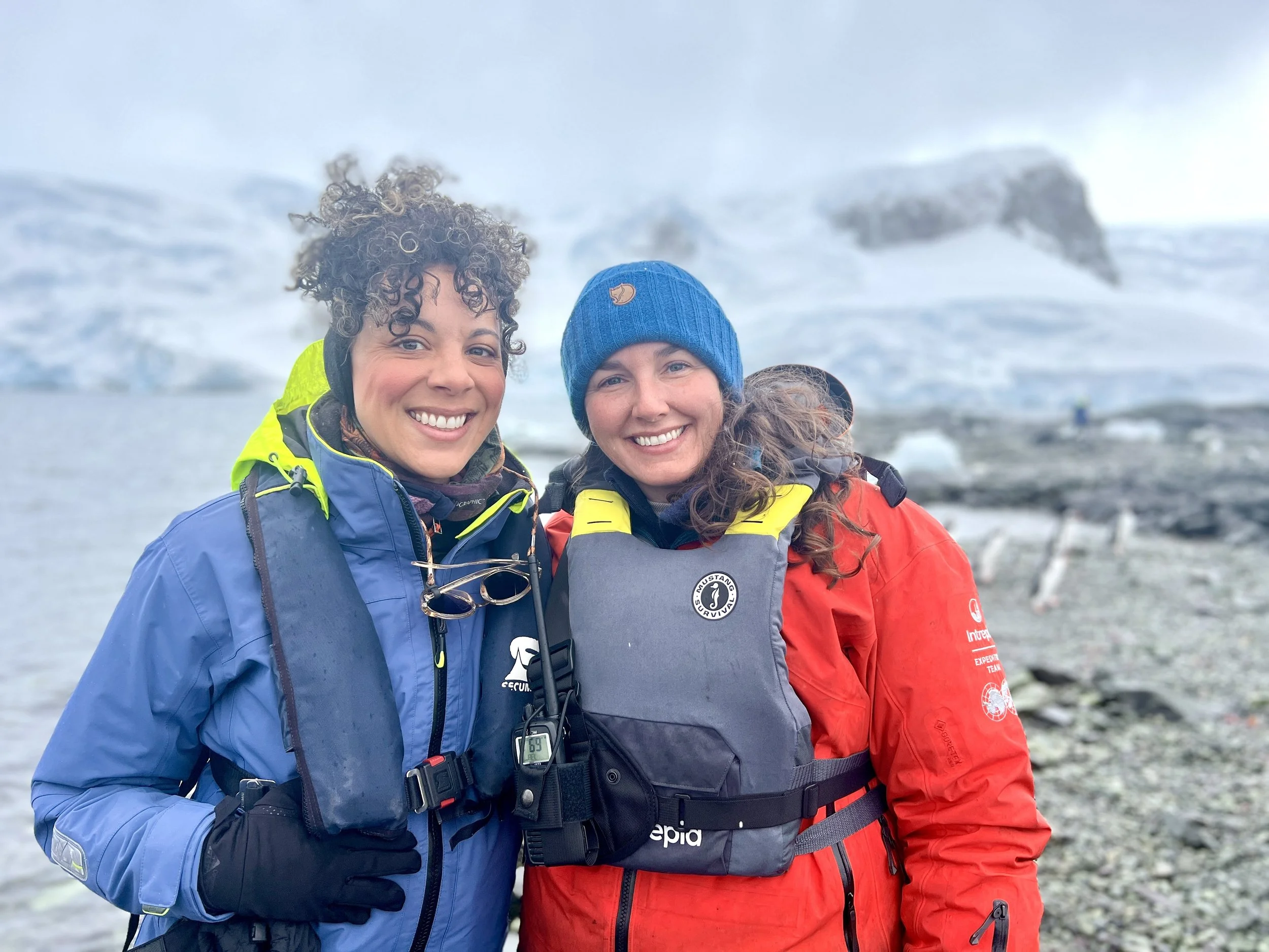 Two women in outdoor gear smiling in a cold, glacier-like environment with ice and rocky terrain, wearing bright jackets and winter hats.