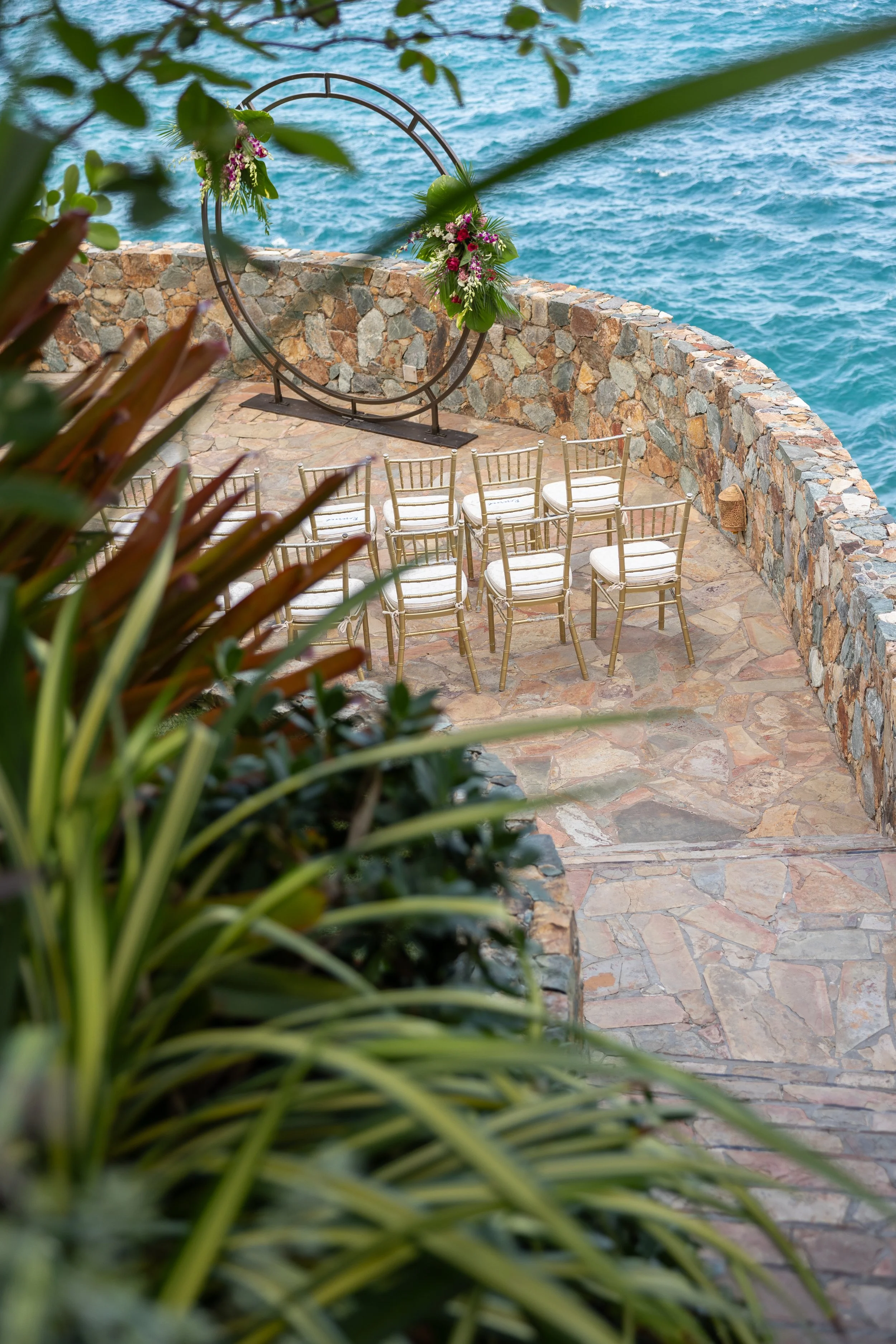Outdoor wedding ceremony setup with eight gold chairs and a large floral ring arch overlooking the ocean, surrounded by plants and stone pavement.