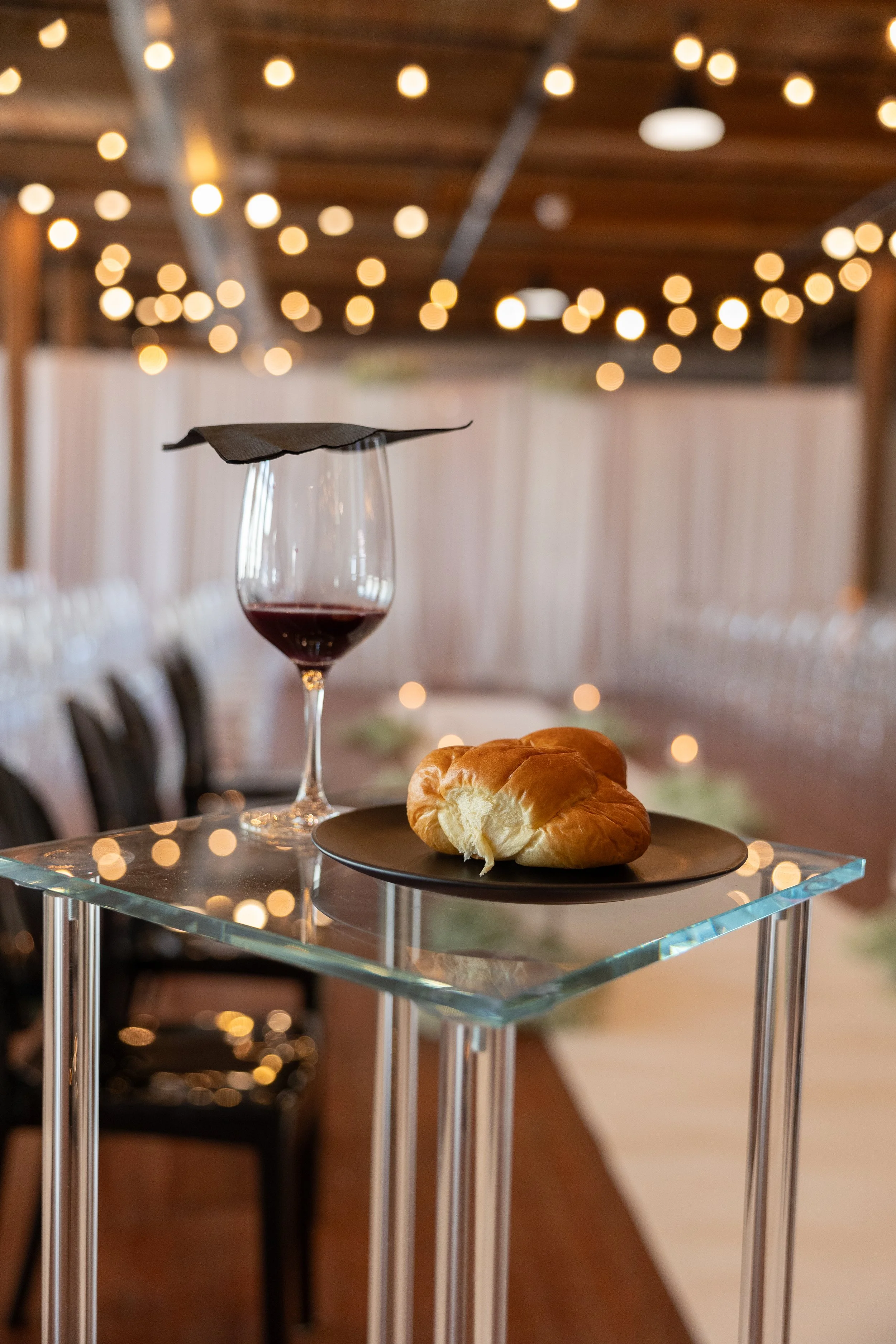 A glass of red wine and a piece of bread on a black plate on a glass table with string lights in the background.