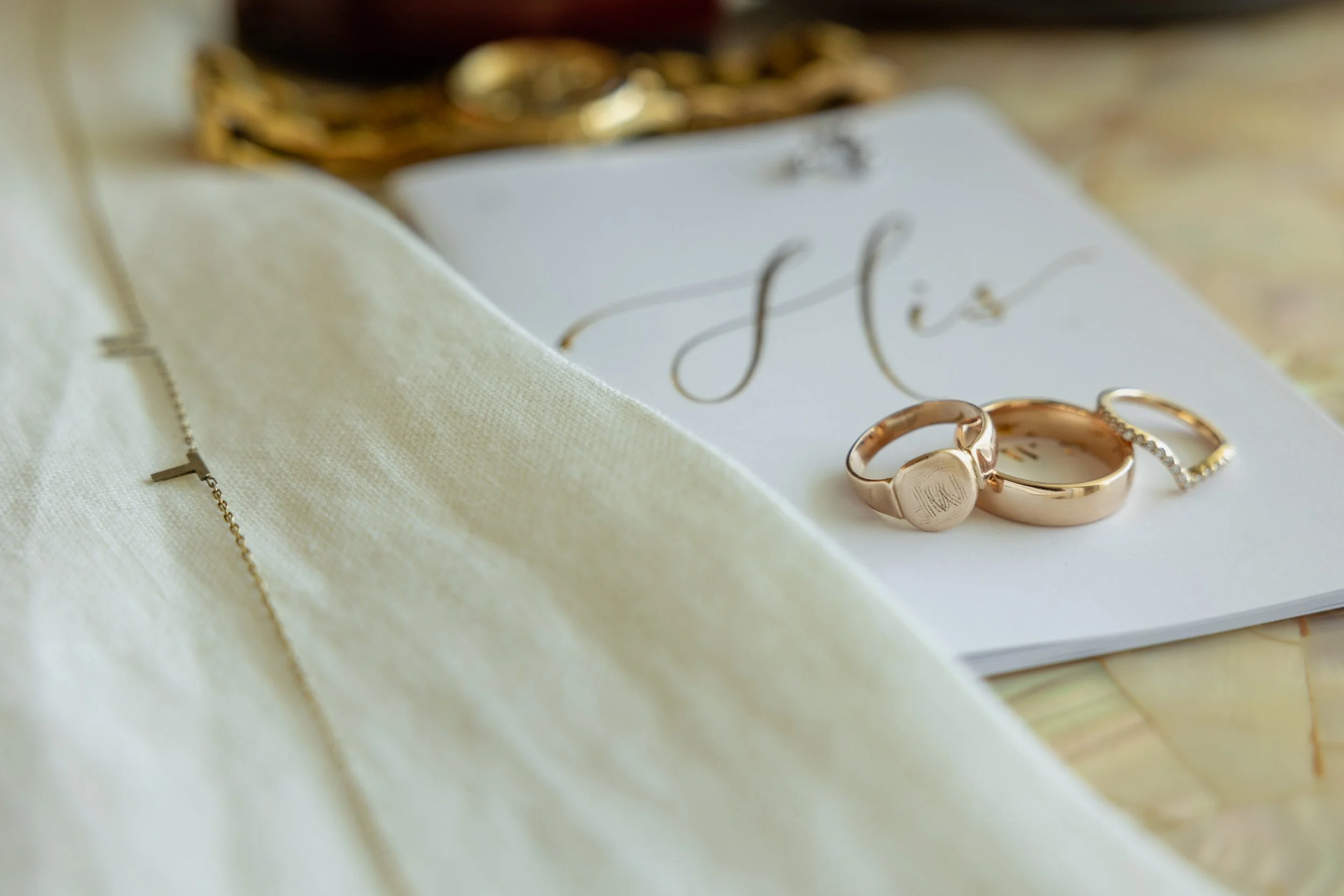 Close-up of wedding rings, a necklace, and a wedding invitation on a marble surface, with a soft fabric in the foreground.