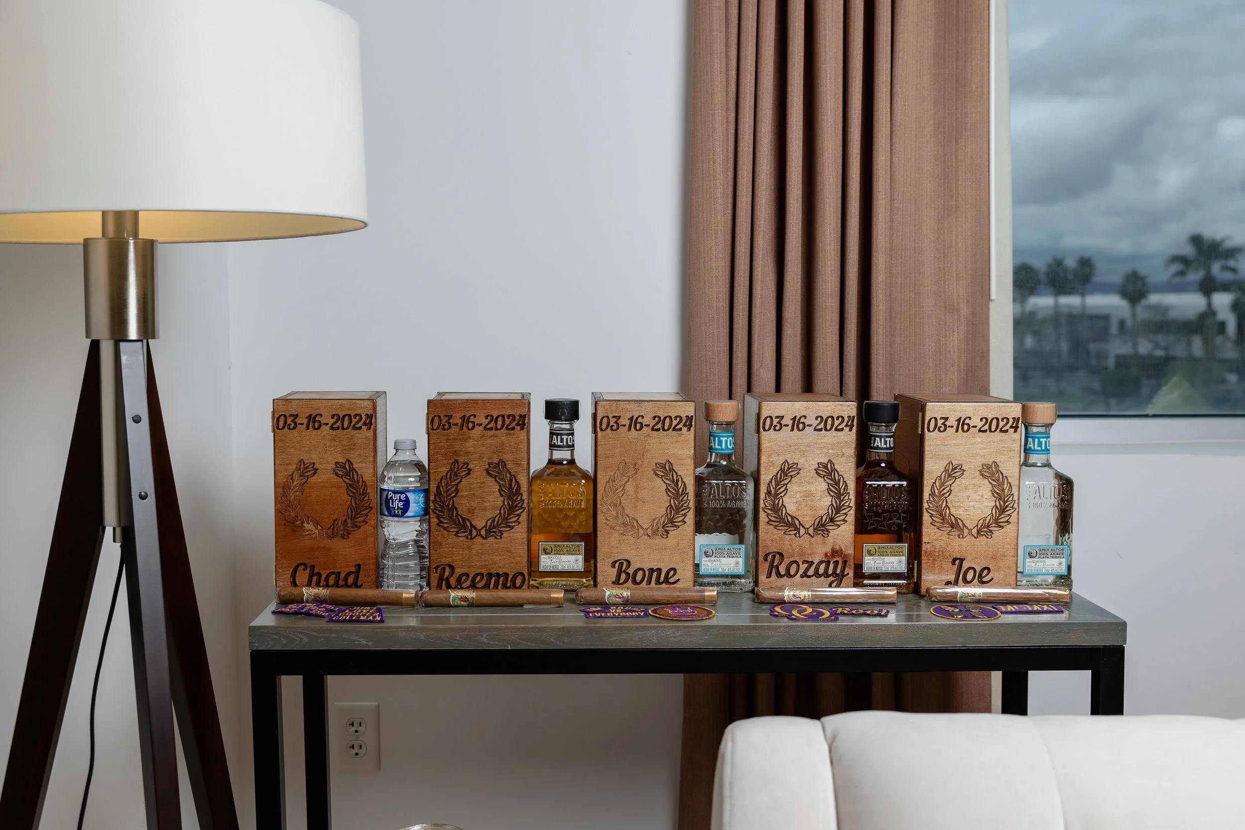 Display of personalized alcohol bottles with wooden boxes and date, name, and color codes, arranged on a table near a window with curtains and a lamp.