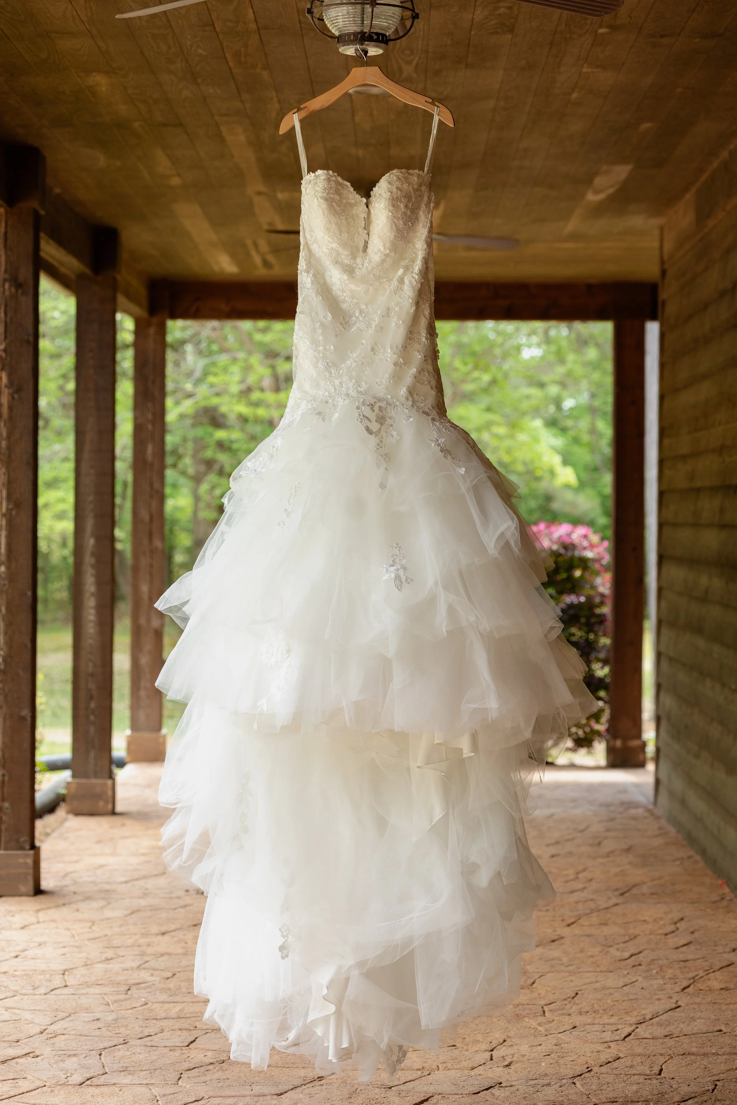 A white wedding dress with lace details and multiple layers of tulle, hanging on a wooden hanger against a wooden porch background.