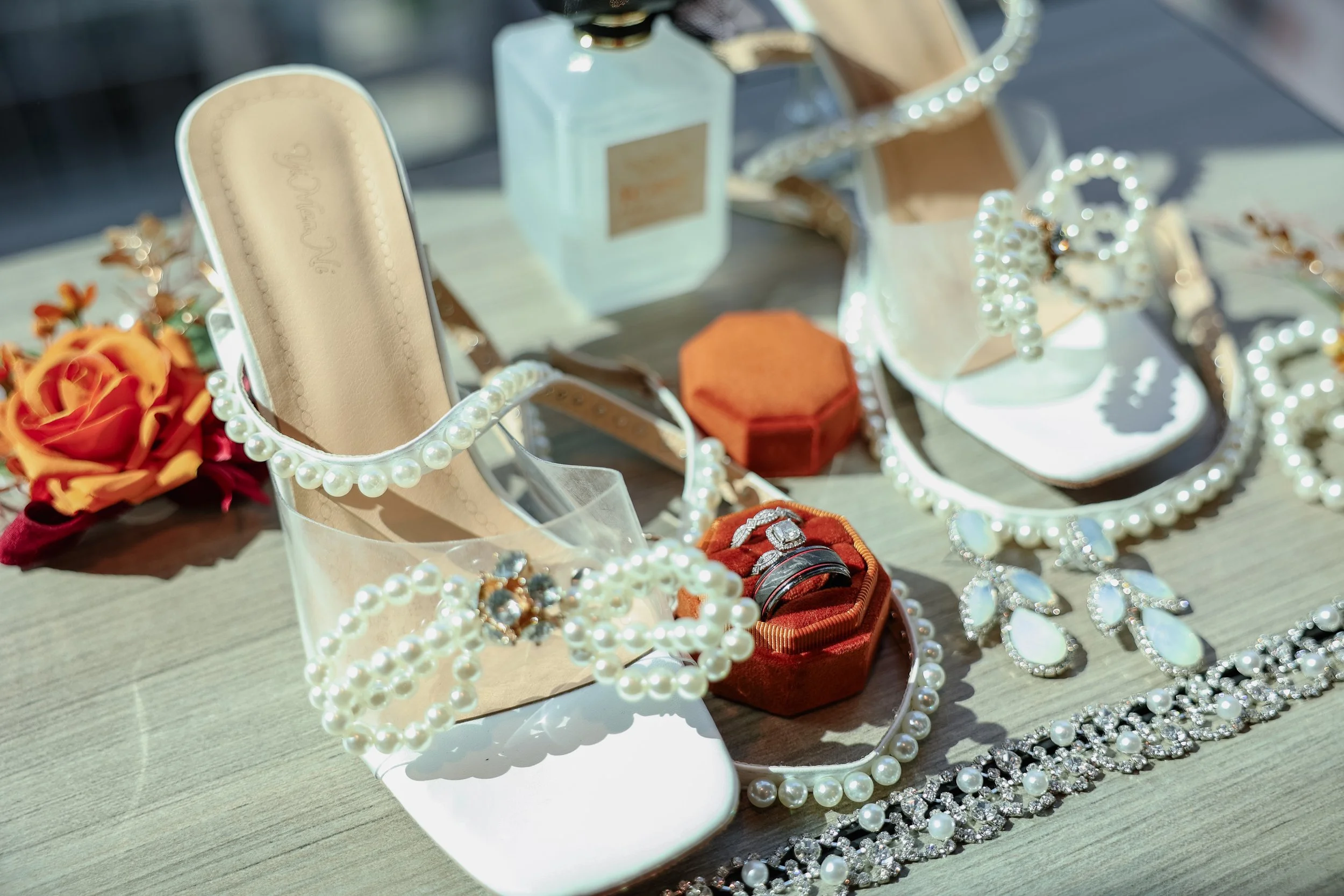 Display of jewelry including pearl necklaces, rings, and earrings, along with shoes, perfume, and floral accessories on a wooden surface.
