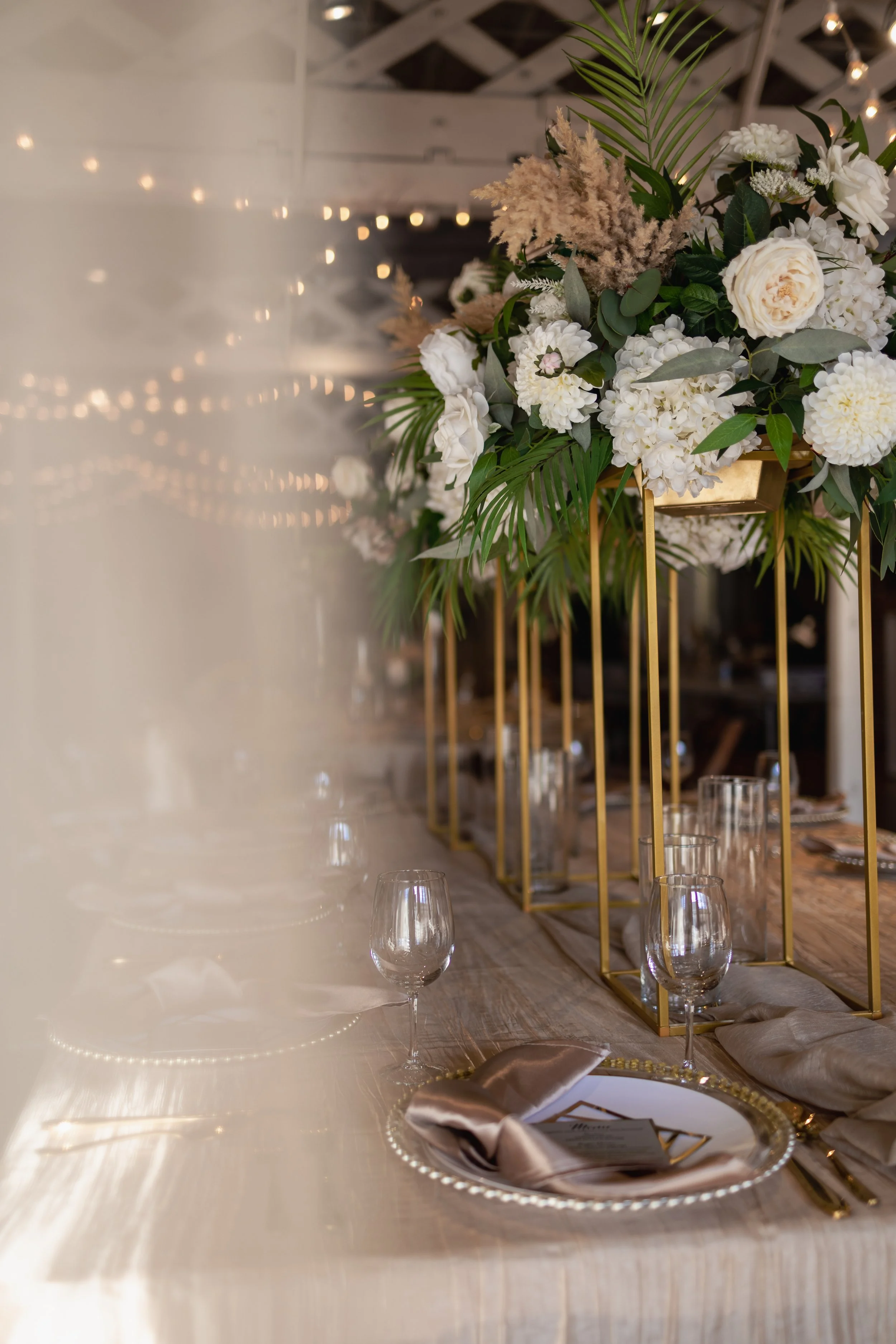 Elegant banquet table with tall gold stands holding white floral centerpieces, set with clear glassware, cream-colored napkins, and a menu on a silver-rimmed plate.