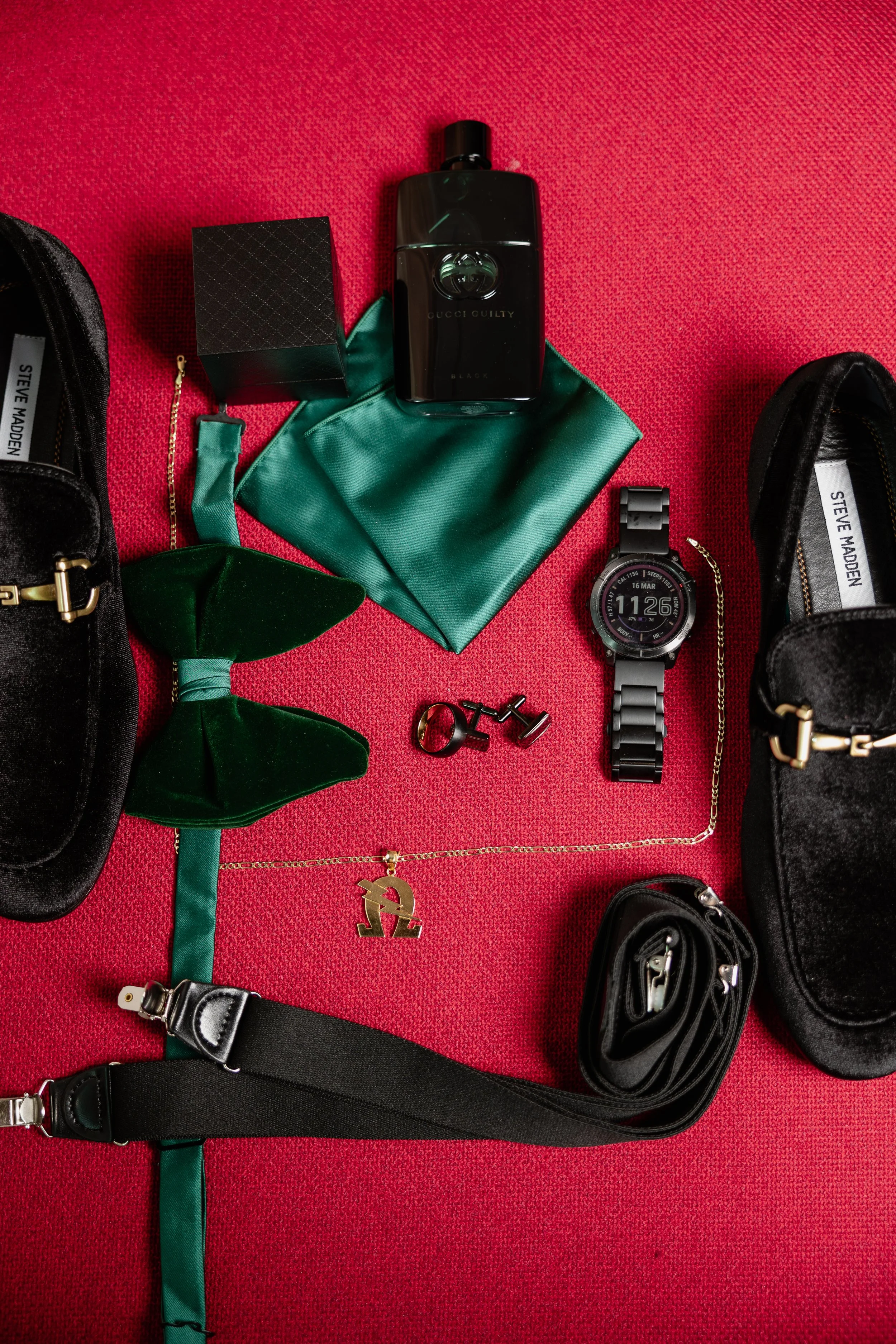 Collection of fashion accessories and personal items on a red surface, including a black Gucci Guilty Black perfume, a black wristwatch, a black strap, gold jewelry, and a black bag.