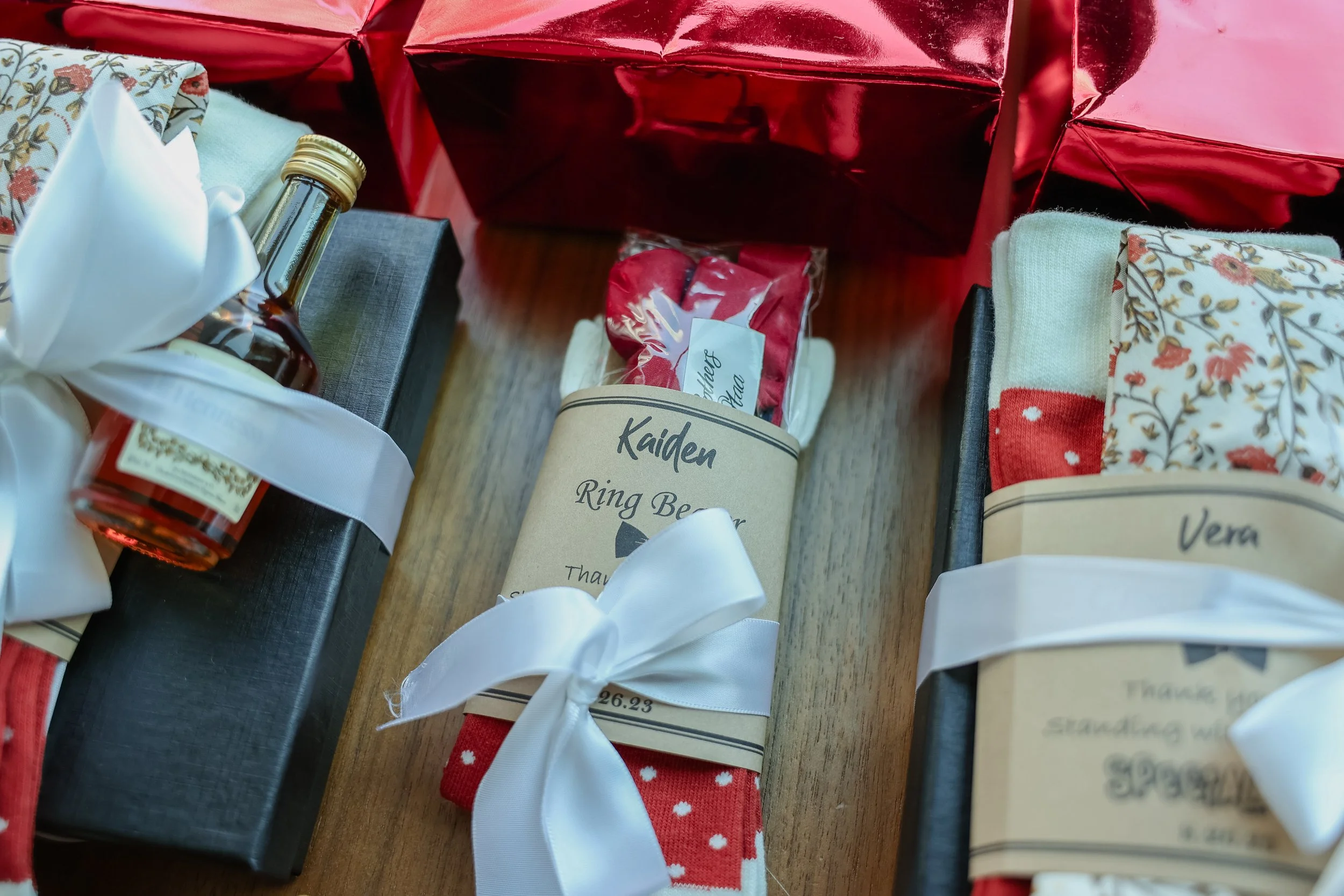 Gift boxes wrapped with black paper, white ribbons, and personalized labels, along with decorated socks, all arranged on a wooden surface with red wrapping paper in the background.