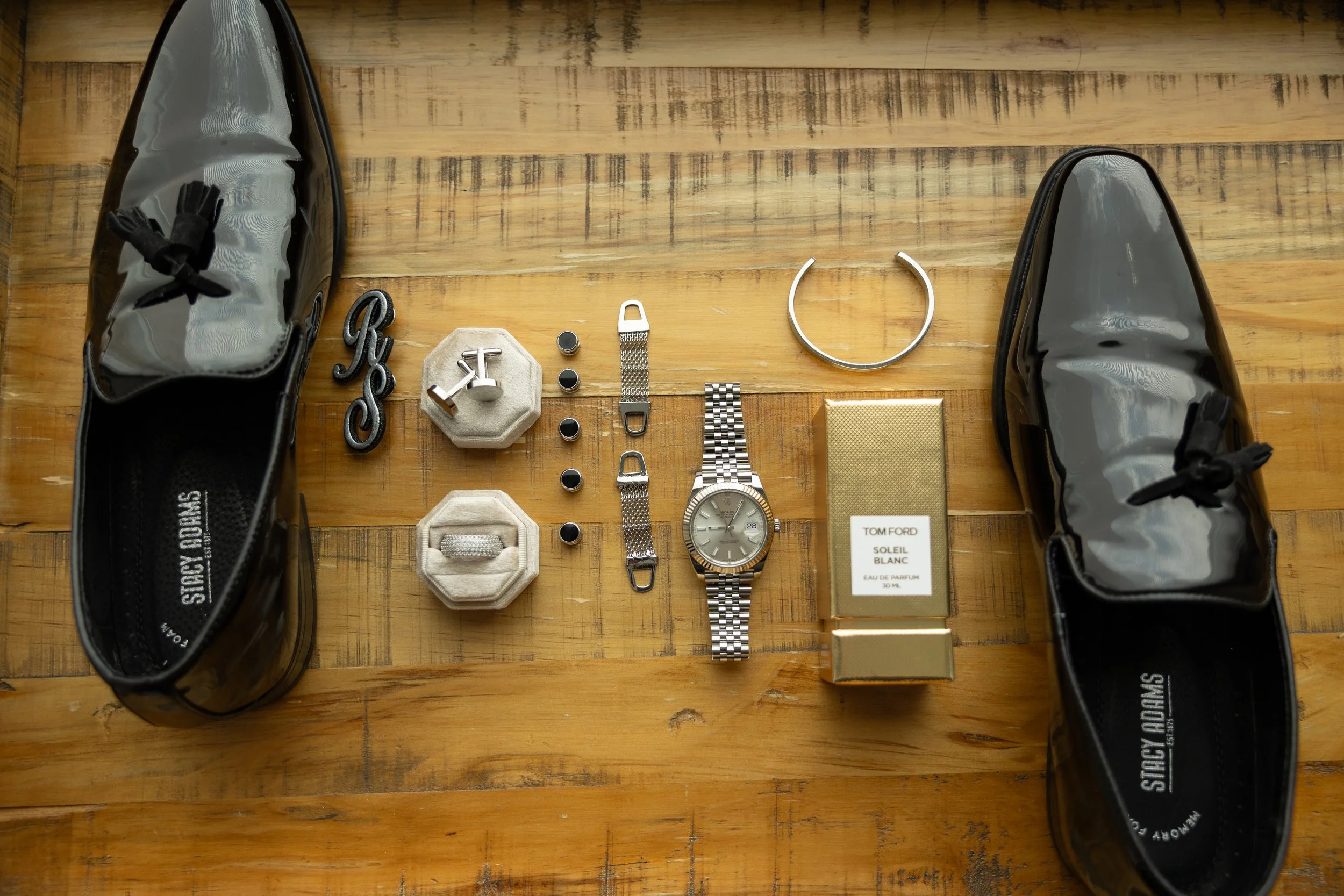 Flat lay of men's accessories and shoes on a wooden surface. Items include black patent leather loafers, black and silver jewelry, a watch, a gold fragrance box titled "Tom Ford Soleil Blanc," and jewelry boxes with rings and cufflinks.