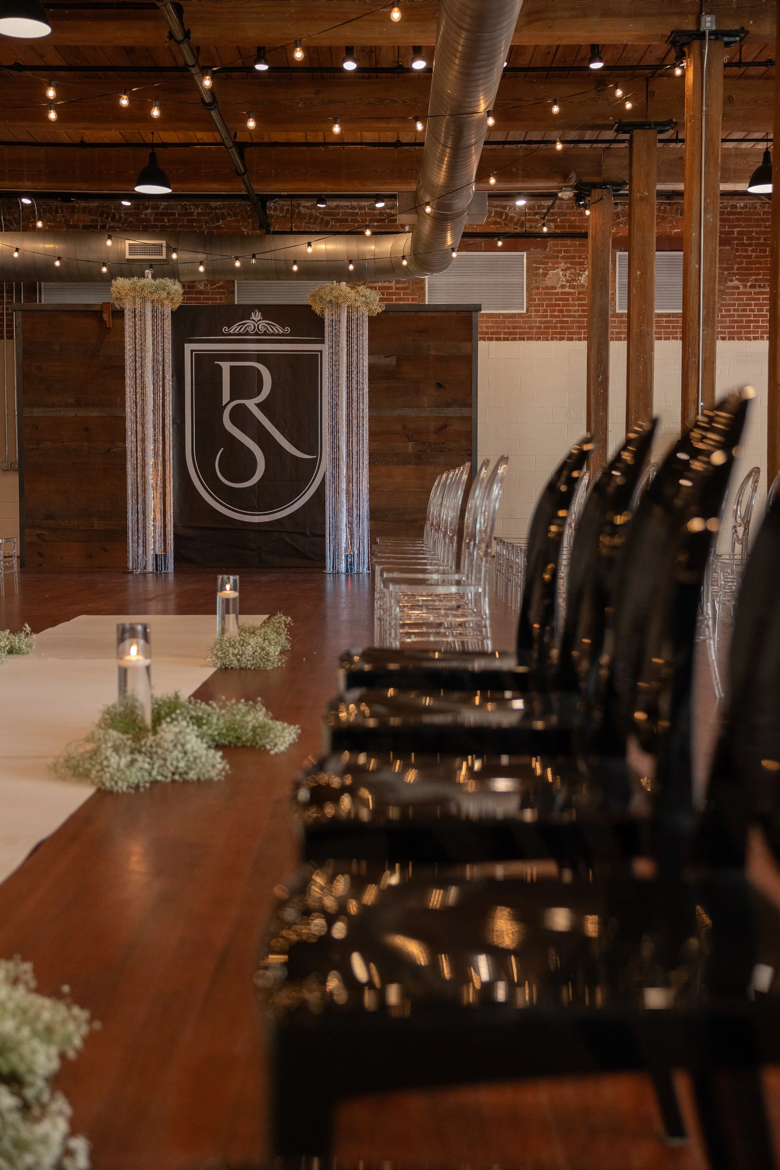 Event space decorated for a wedding, featuring a long wooden table with floral centerpieces and candles, a stage with a monogram backdrop, and hanging string lights, inside an industrial-style venue with brick walls and wooden beams.