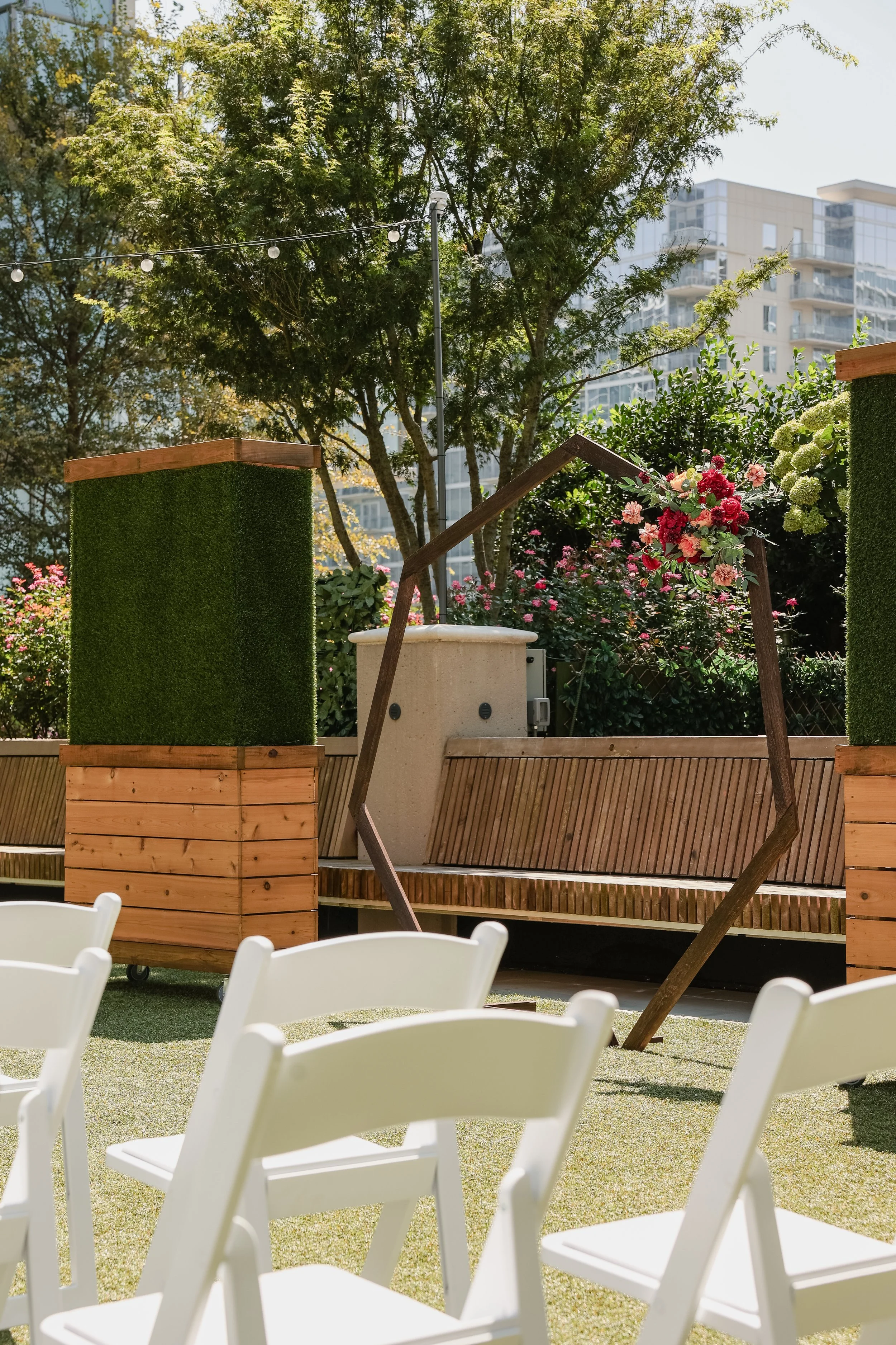 Outdoor event setup with white folding chairs on a grassy lawn, flower arrangements on wooden planters, a wooden bench, decorative string lights, lush green trees, and modern high-rise buildings in the background.