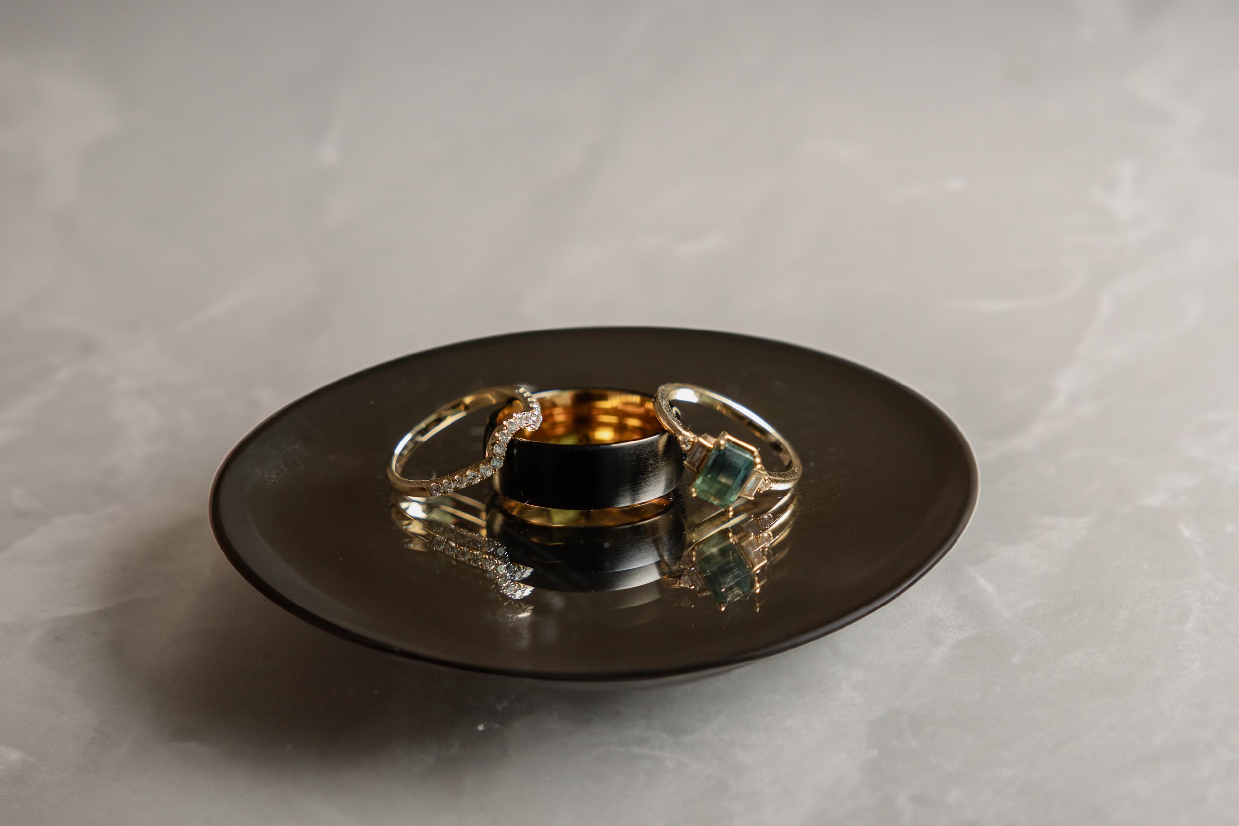 A black dish with three wedding rings on it, including a diamond-studded band and a green gemstone ring, on a marble surface.