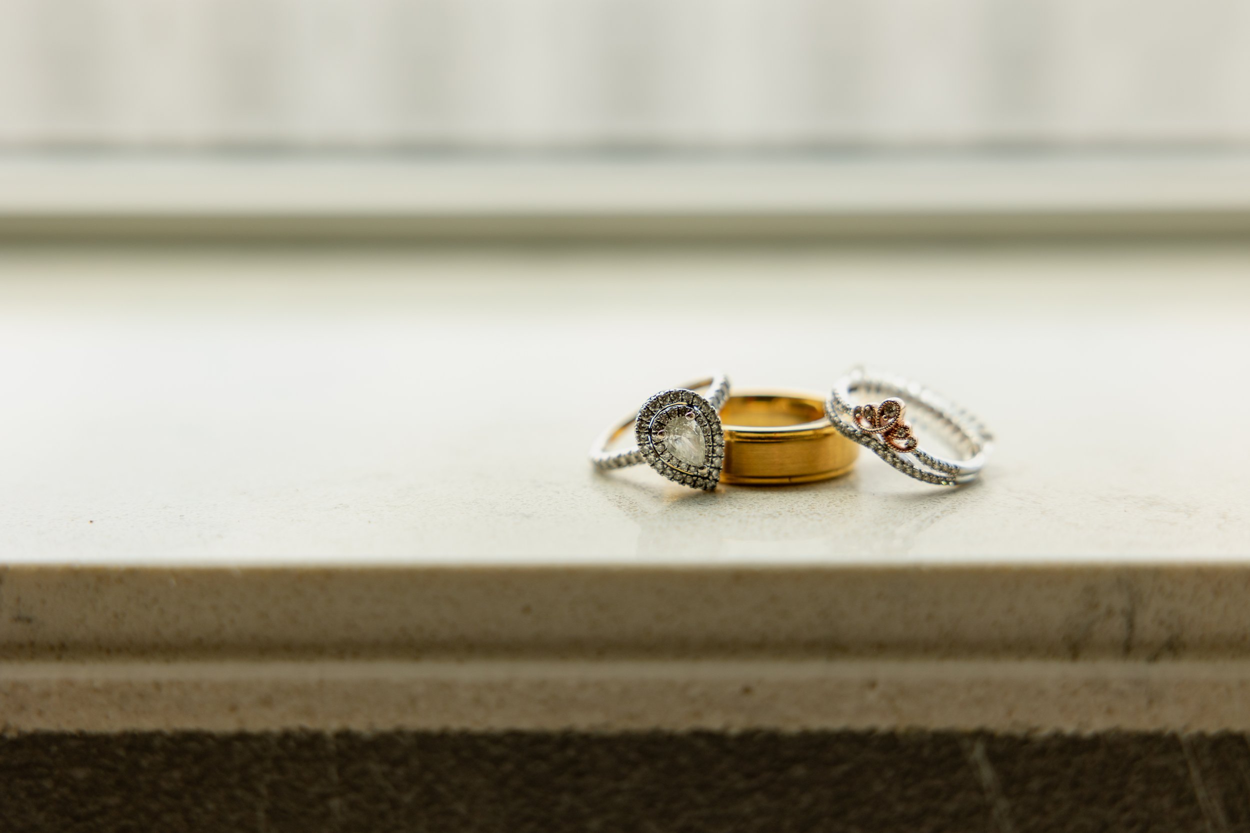 Three rings, two silver and one gold, placed on a light-colored surface with a window or light source in the background.
