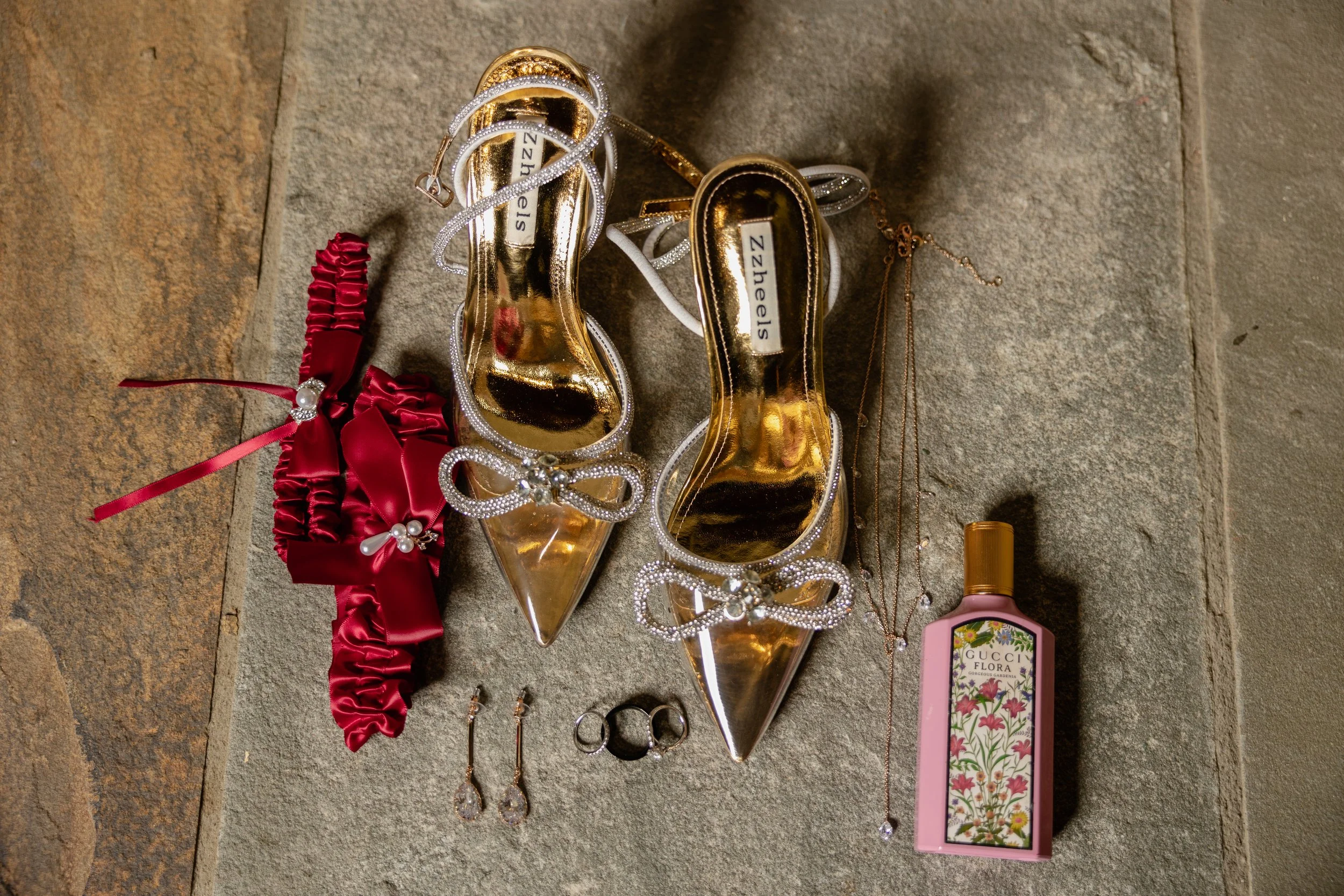 Gold high-heeled shoes with rhinestone bows, a red satin garter with pearl accents, jewelry including earrings, rings, and layered necklaces, and a pink bottle of Gucci Flora perfume on a textured surface.