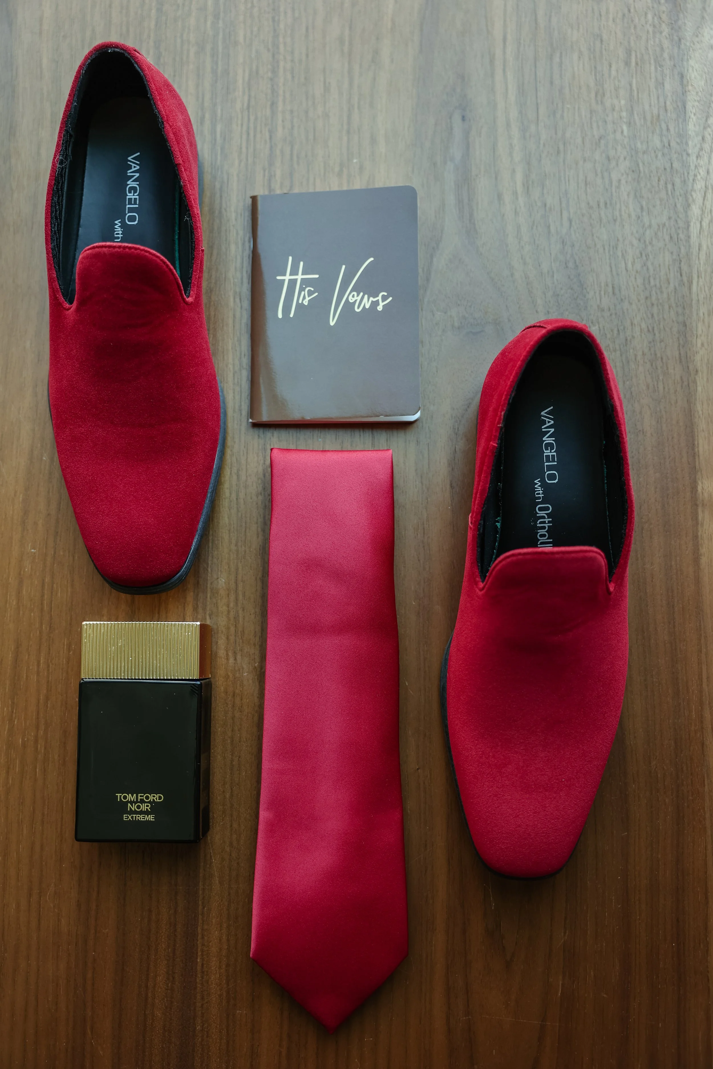 A red pair of shoes, a black and gold box of Tom Ford perfume, a red pocket square, a red tie, and a small notebook with the words 'His Vows' on the cover are arranged on a wooden surface.