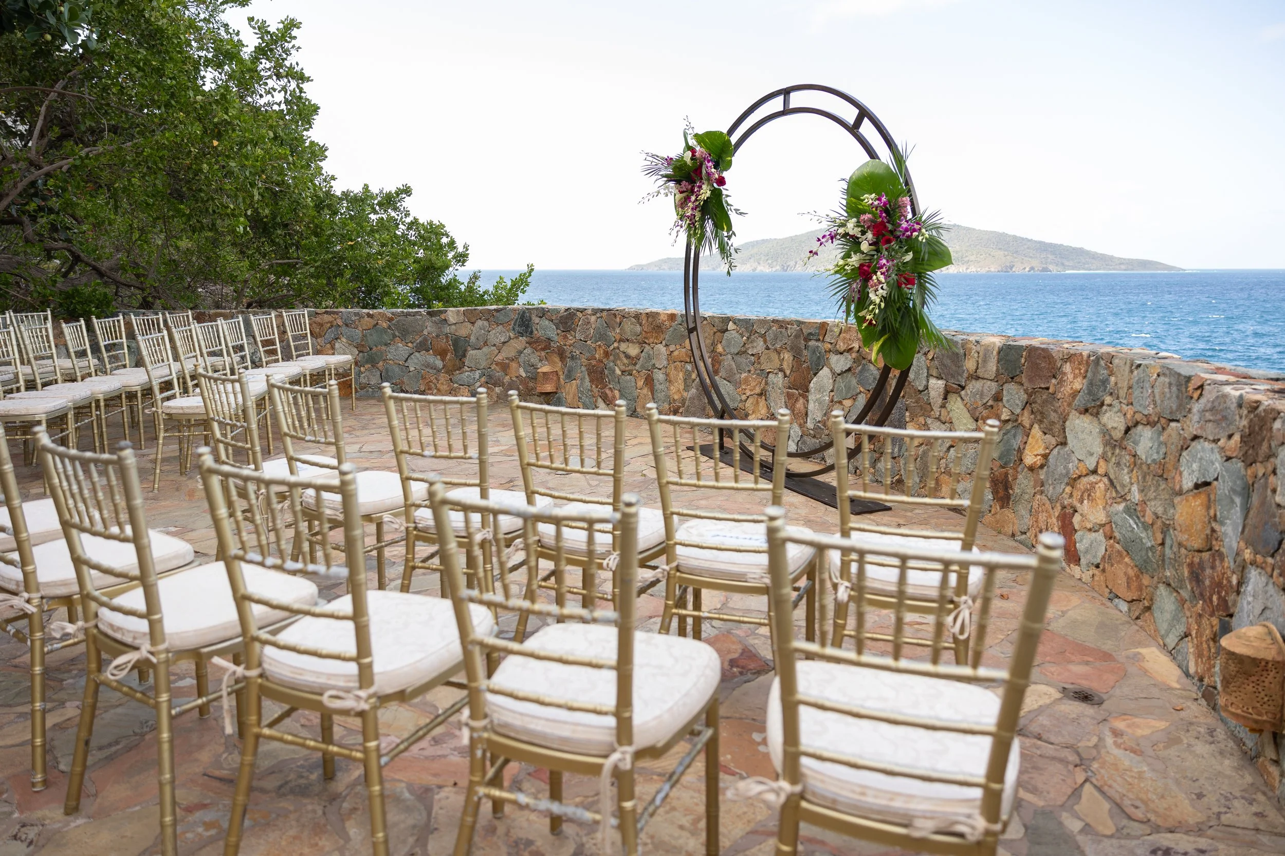 Outdoor wedding setup with gold chairs arranged in rows, a circular floral arch, and ocean view in the background.