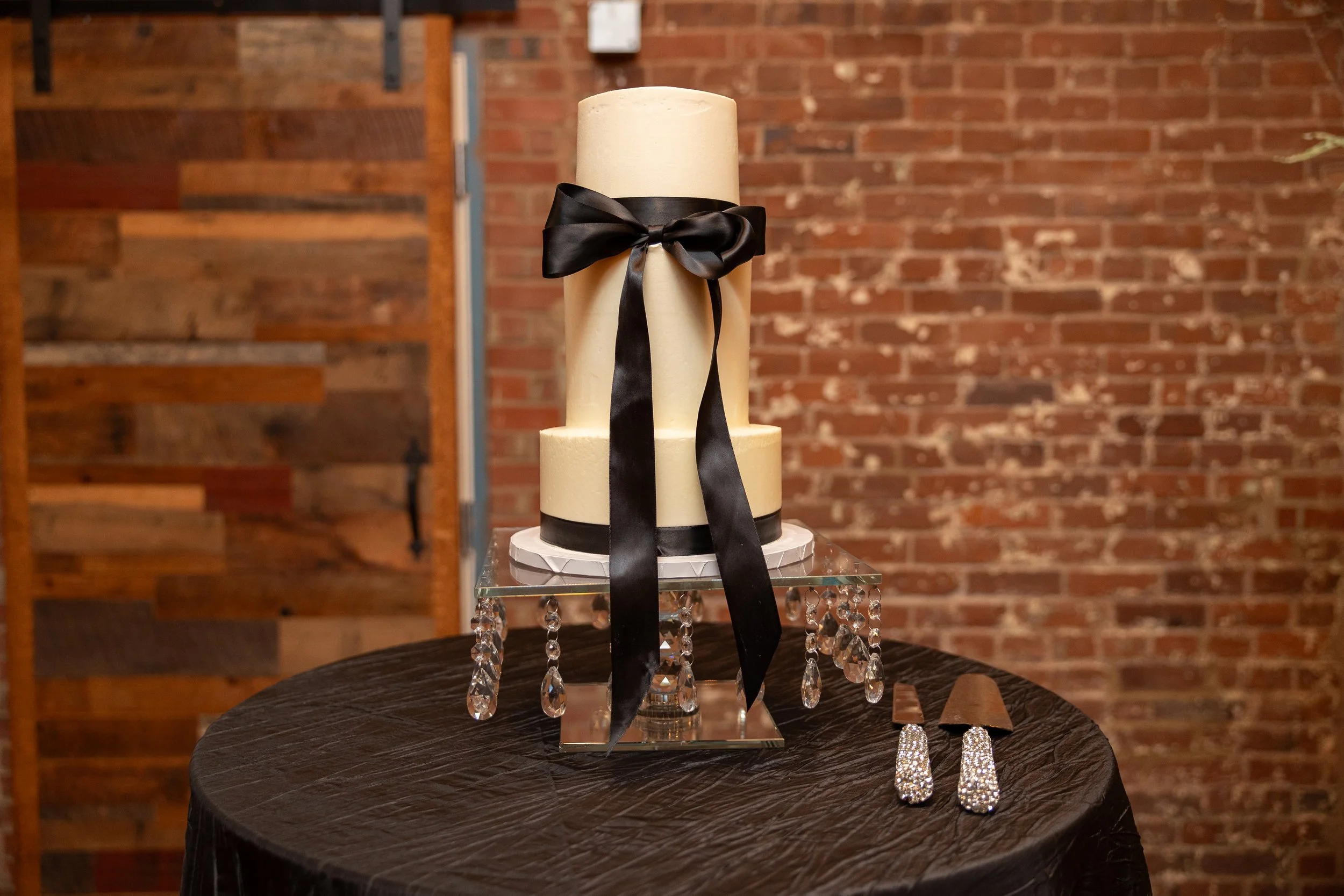 A two-tiered white wedding cake decorated with a black satin ribbon bow on top, displayed on a glass stand with hanging crystal ornaments. Next to it are a pair of sparkly chandelier earrings and a matching sparkly hair clip on a black tablecloth aga