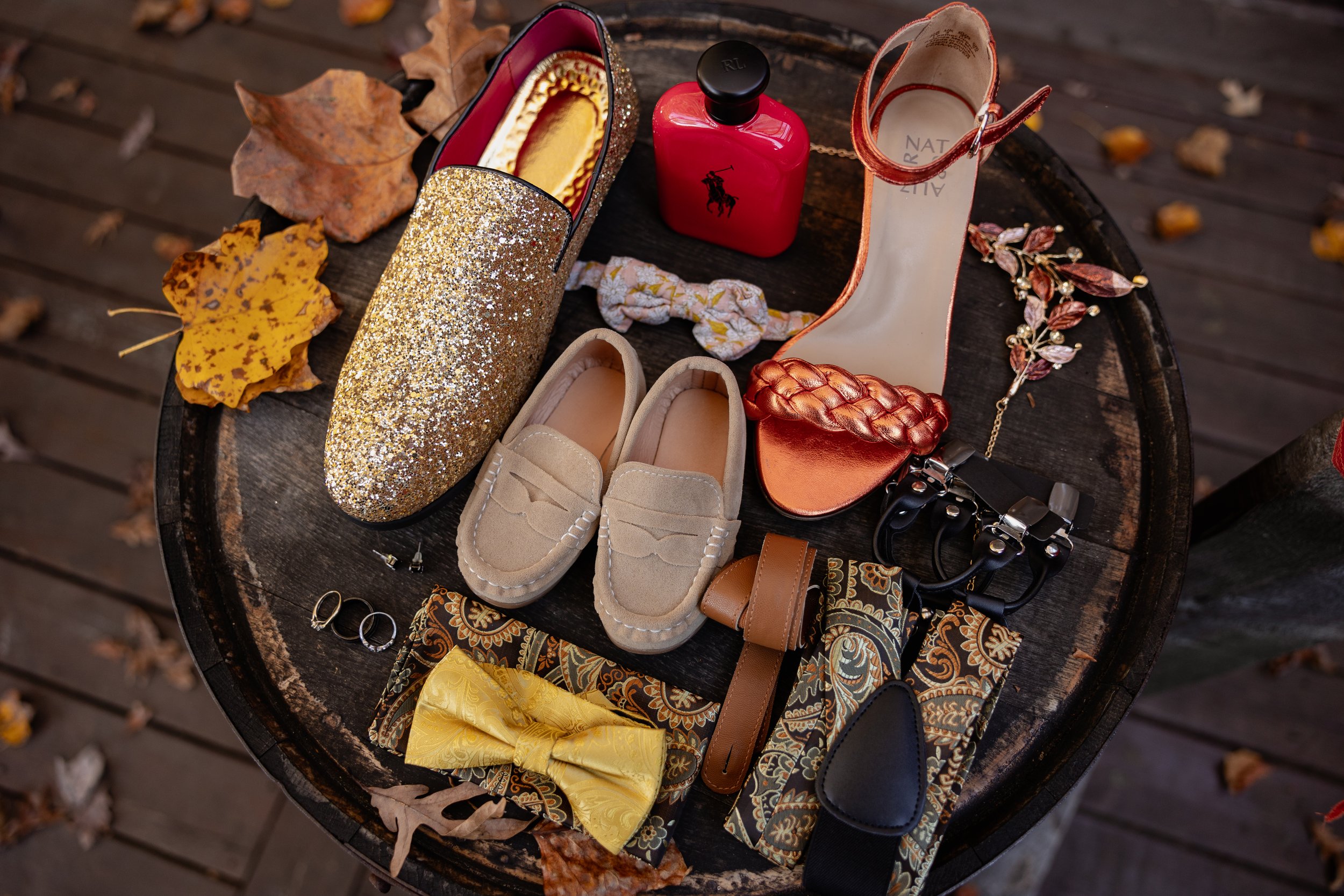 A collection of women's accessories and shoes on a wooden surface, including a glittery gold slip-on shoe, beige loafers, an orange satin heel with braided detail, a printed scarf, a yellow bow, a brown belt, rings, a black hair scrunchie, a floral h