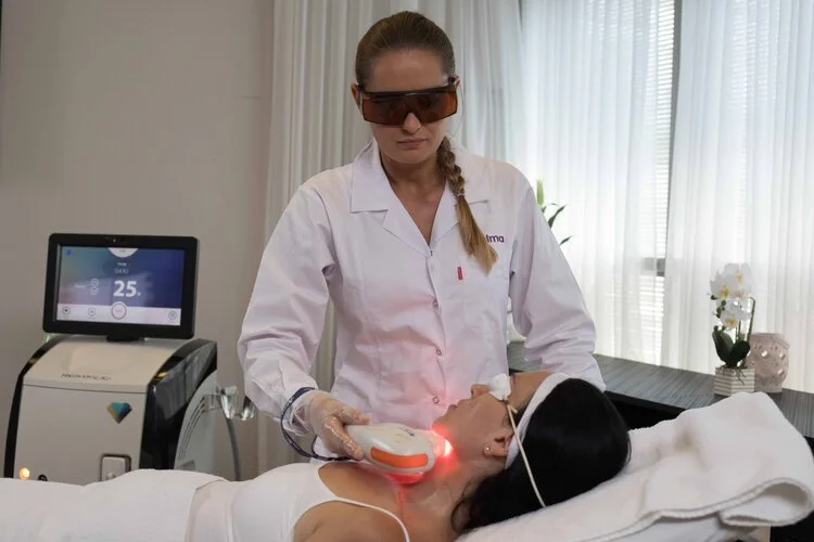 A beauty technician wearing protective goggles performs a laser treatment on a woman's neck using a handheld device in a clinic setting.