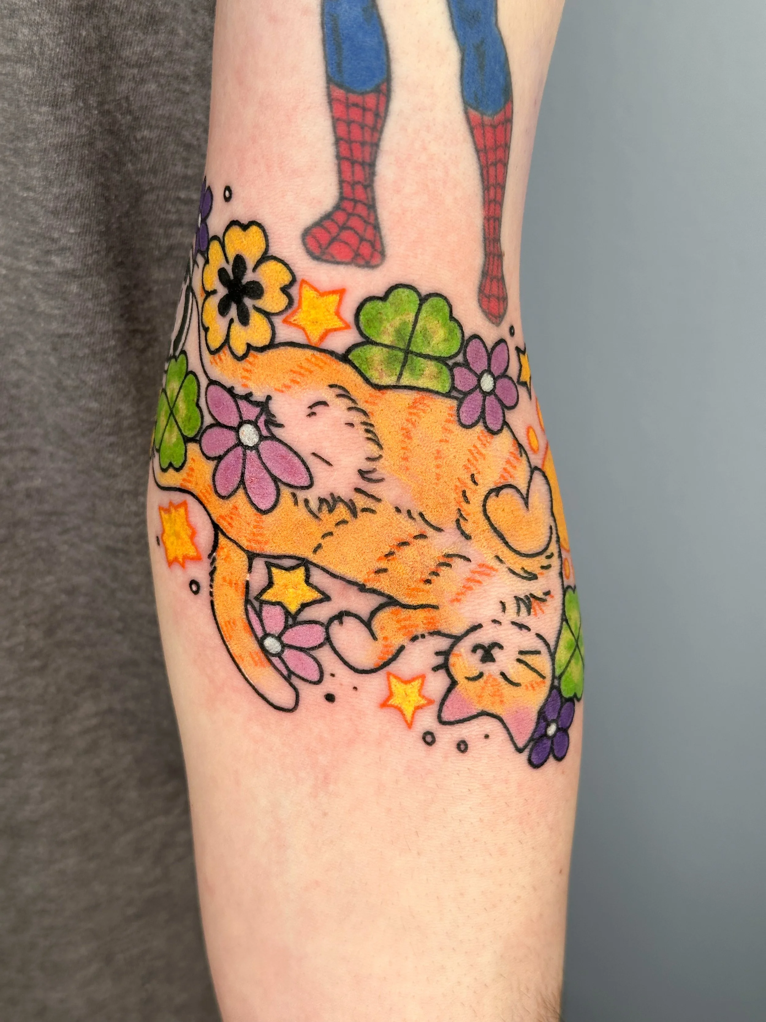 Colorful tattoo on forearm featuring a sleeping kitten surrounded by purple and yellow flowers, green leaves, and stars.
