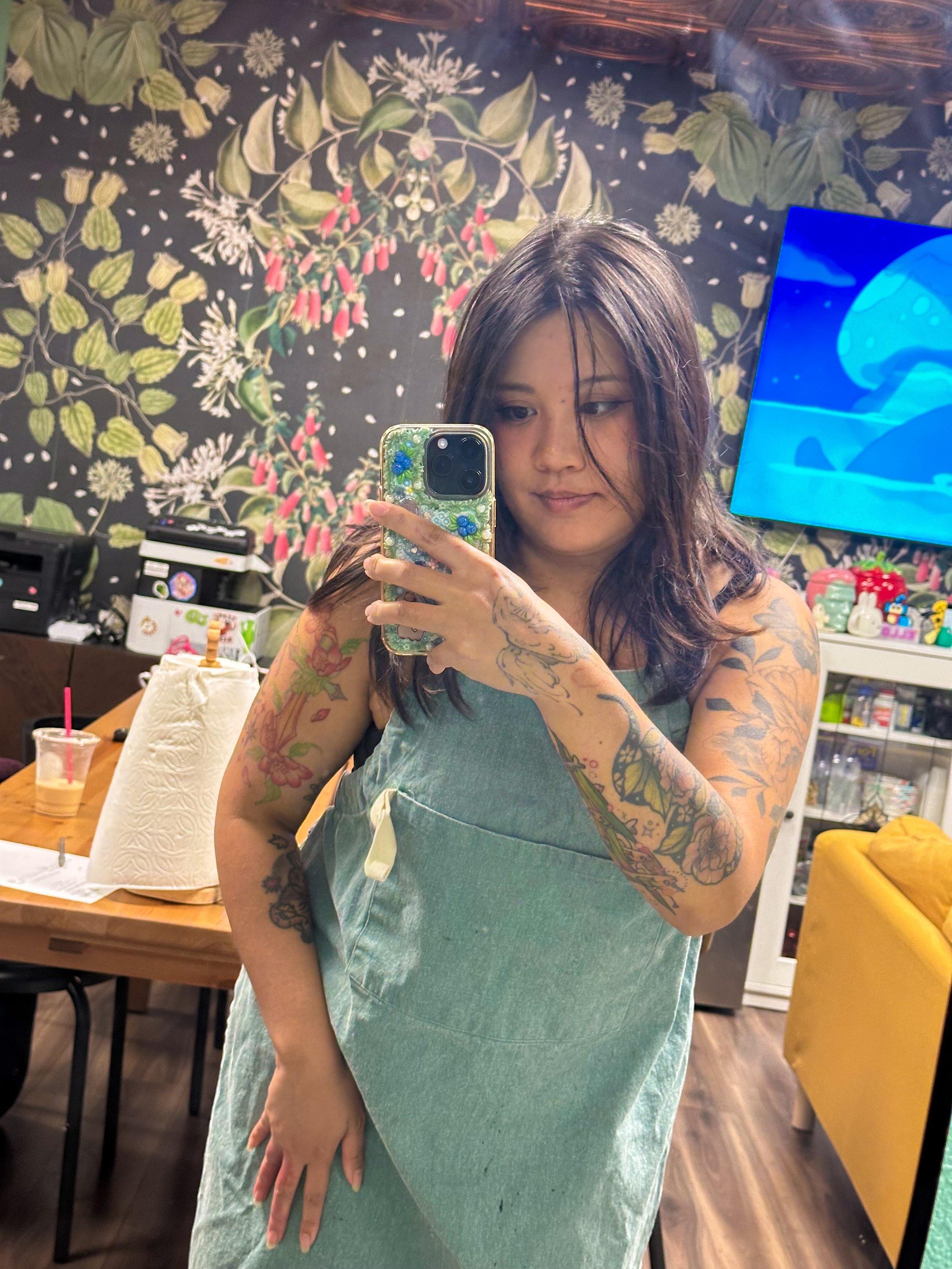 Young woman with shoulder-length dark hair and tattoos on her arms taking a mirror selfie, wearing a sleeveless dress in a room with a floral mural on the wall, a TV screen, a wooden table with a paper towel roll and a drink, and a yellow couch.