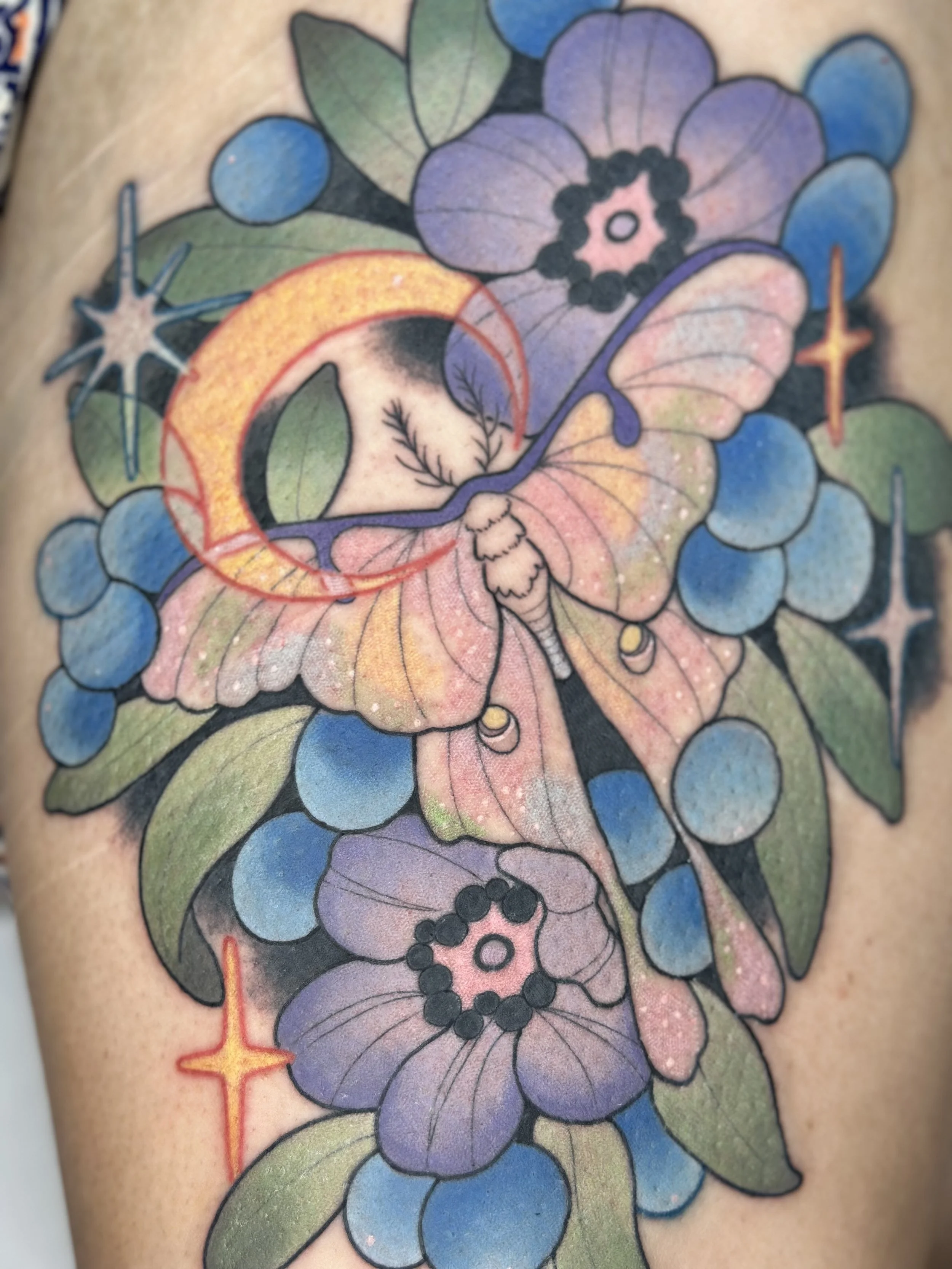 Colorful tattoo featuring a butterfly with large purple wings, surrounded by blue flowers, green leaves, and star-like symbols with orange and red accents.