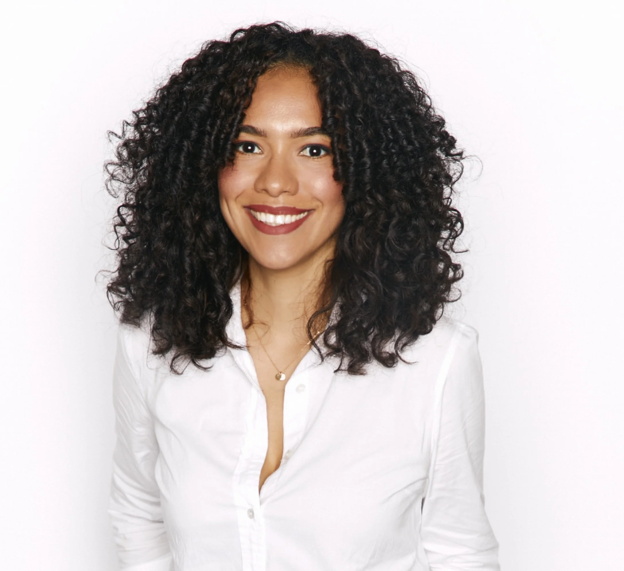 Portrait of a woman with dark, curly hair, wearing a white shirt, smiling, against a white background.