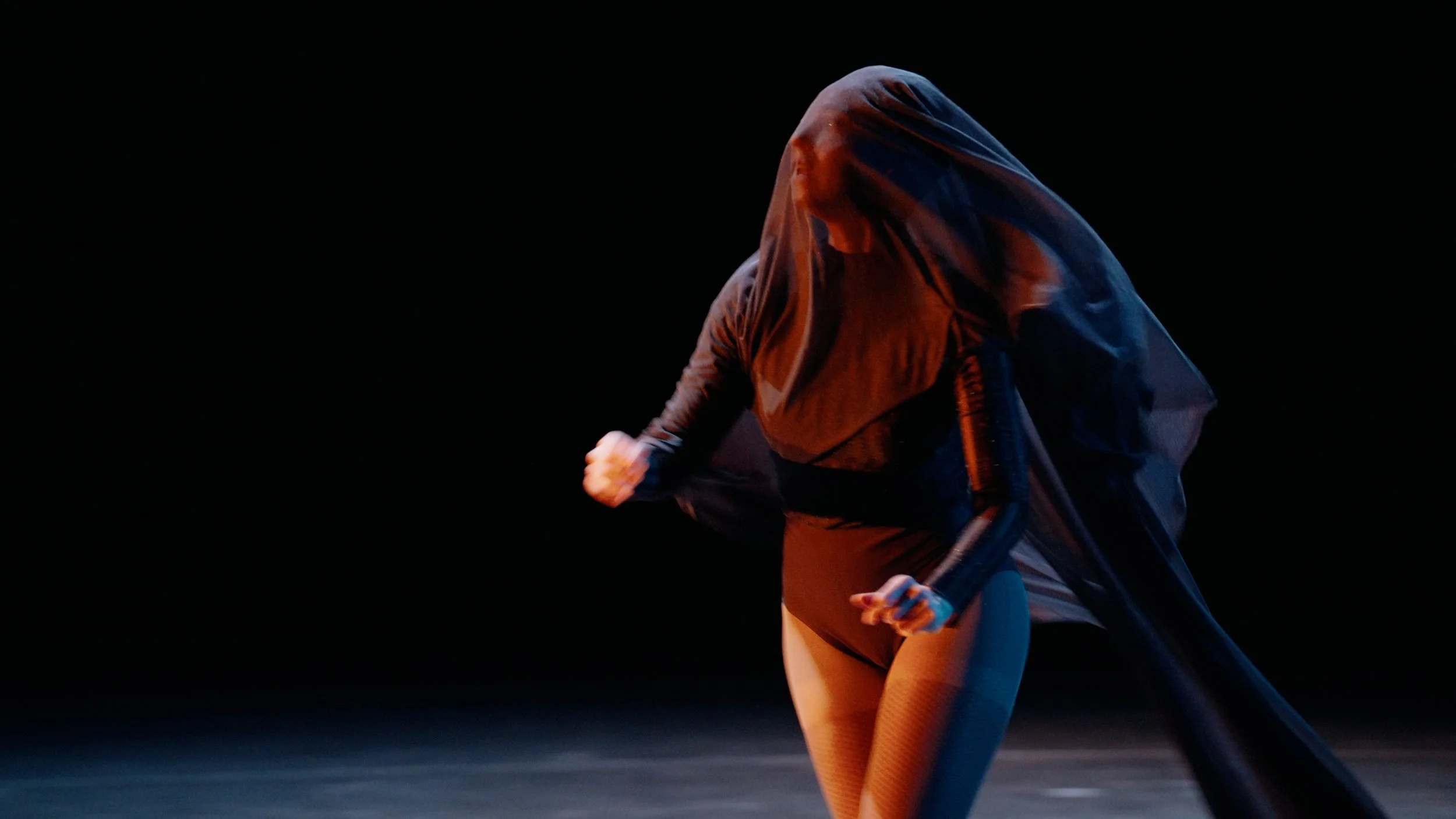 Dancer in a dark studio wearing a sheer black top and shorts, with lighting that creates a dramatic shadow effect.